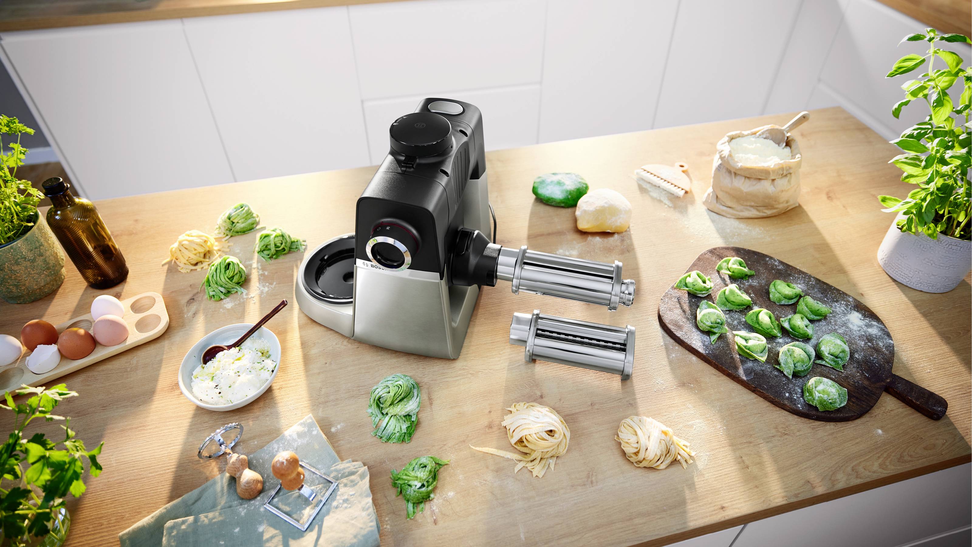 Machine à pâtes sur un plan de travail de cuisine avec des pâtes fraîchement préparées vertes et jaunes, des œufs et des herbes, suggérant une cuisine maison.