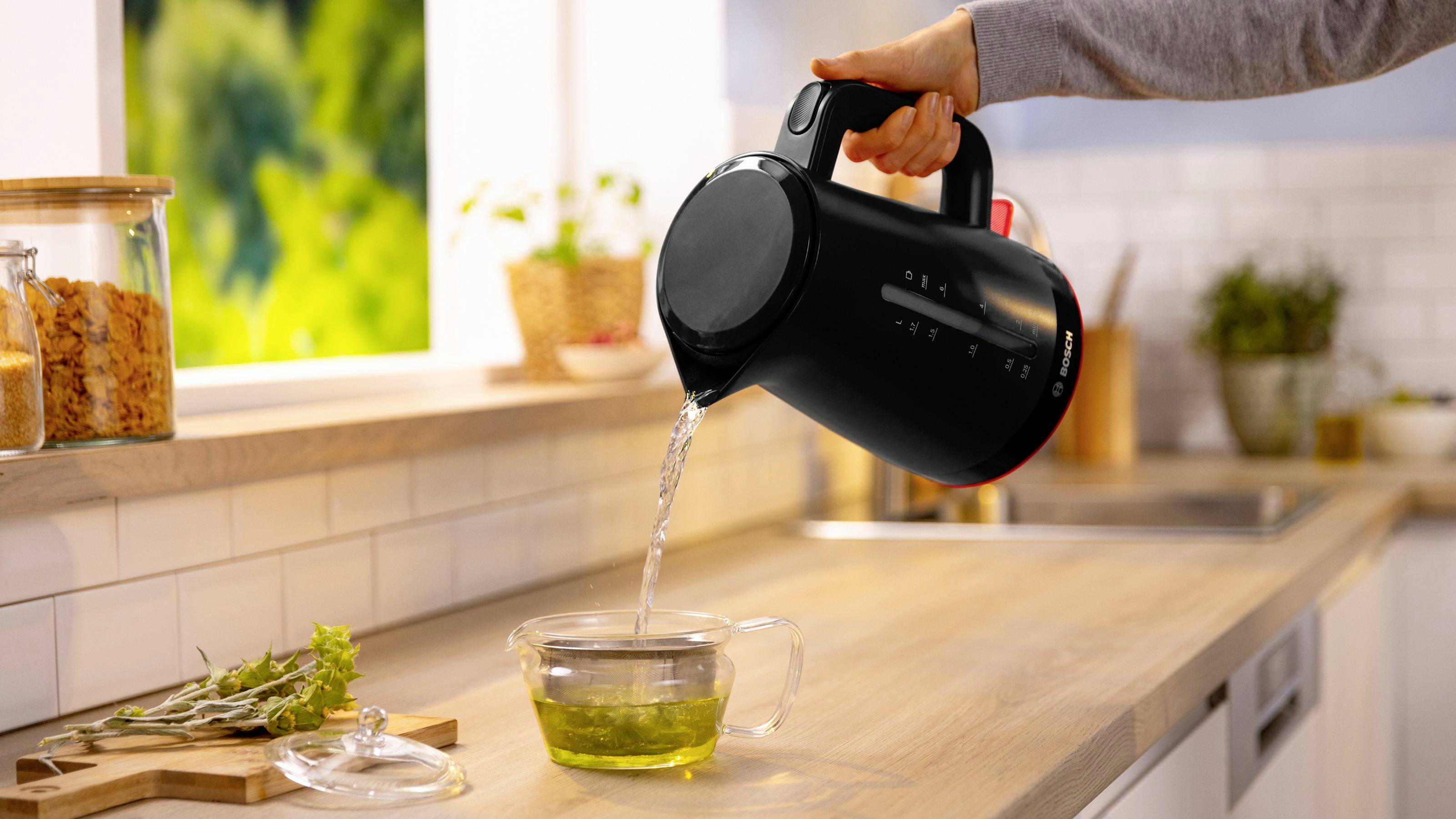 Une personne verse de l'eau chaude d'une bouilloire électrique noire dans une tasse en verre sur un plan de travail de cuisine, avec des plantes et des bocaux en arrière-plan.
