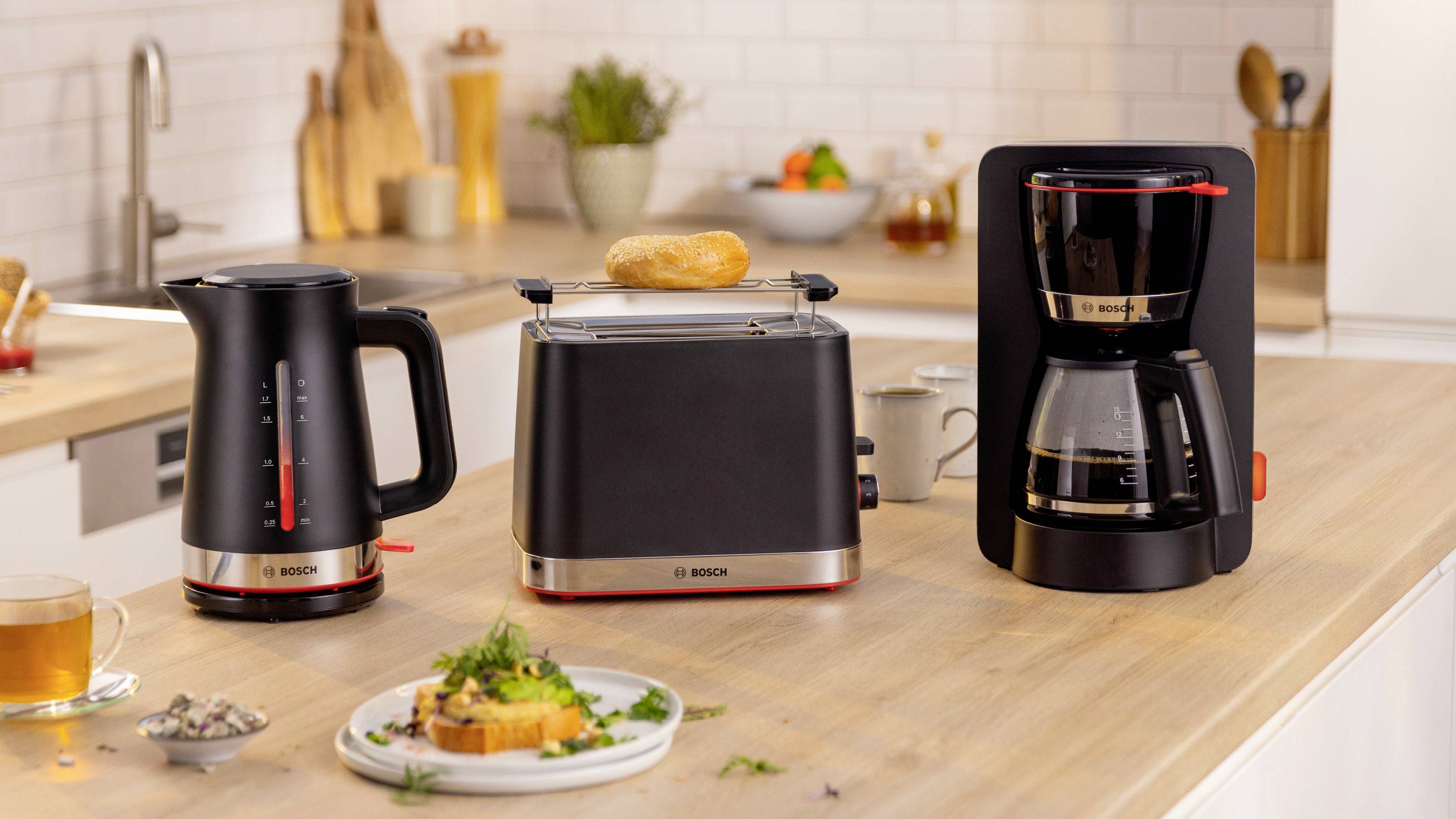 Une cuisine avec une bouilloire, un grille-pain et une cafetière sur un plan de travail. Une tranche de pain est dans le grille-pain. Des aliments et des plantes sont en arrière-plan.
