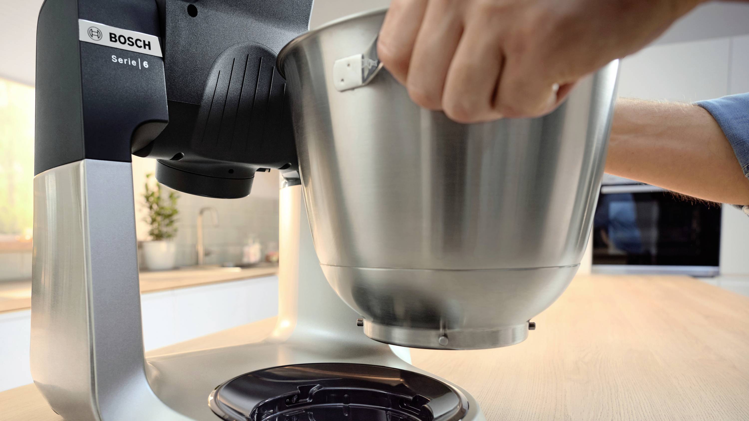 Une personne fixe un bol à mélanger à un mixeur de cuisine Bosch sur un plan de travail en bois dans un environnement de cuisine moderne.