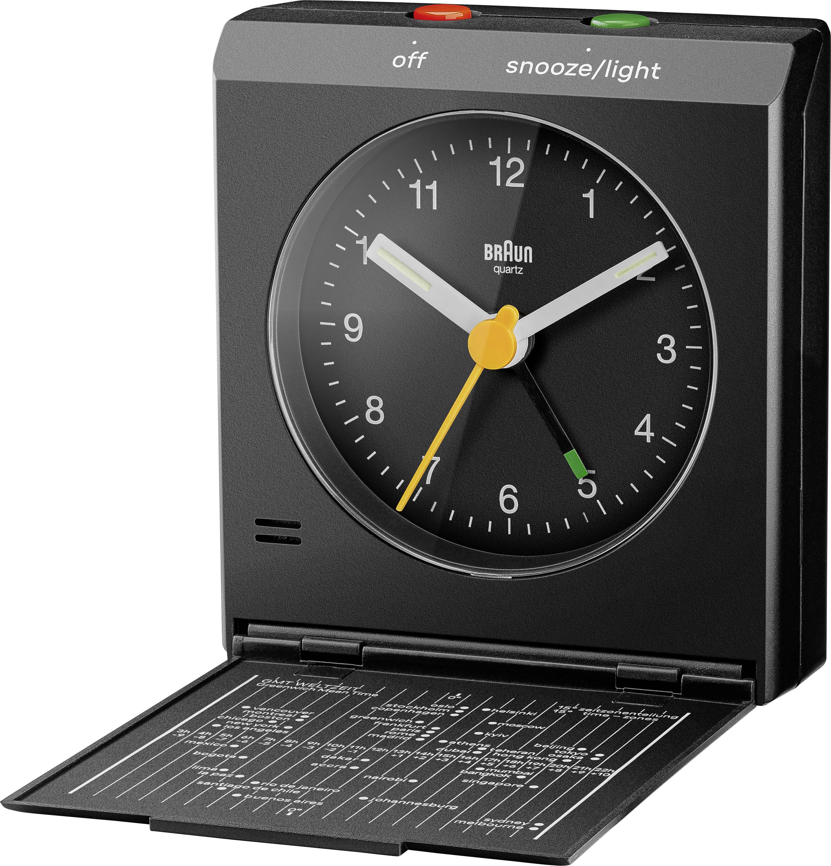 Une horloge de réveil analogique Braun noire avec des aiguilles blanches et jaunes, accompagnée d'un étui de voyage ouvert, révélant un graphique des fuseaux horaires mondiaux à l'intérieur du couvercle.