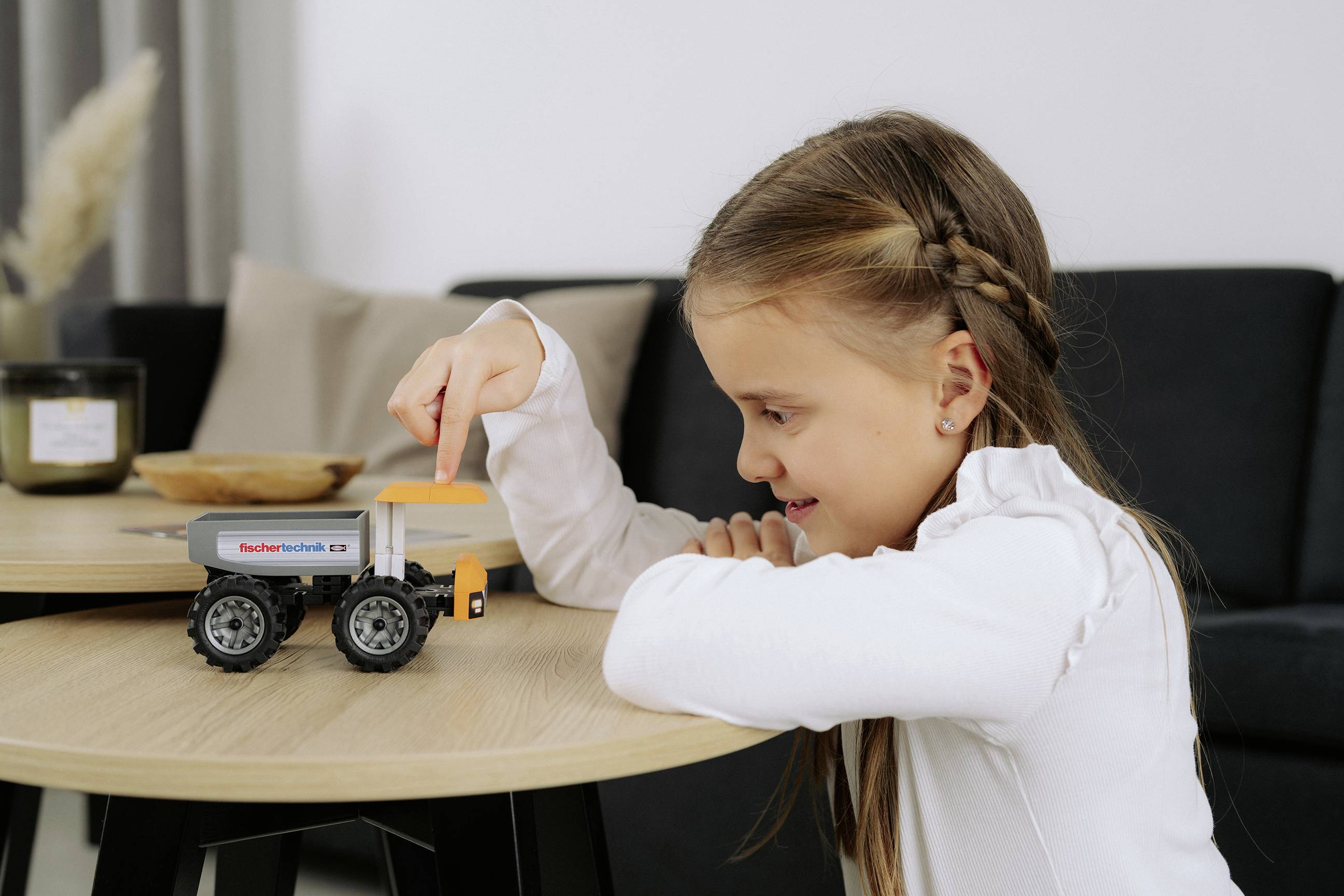 Un enfant jouant avec un camion miniature sur une table en bois, concentré avec intensité. La pièce est doucement éclairée, avec un canapé noir et des objets décoratifs en arrière-plan.
