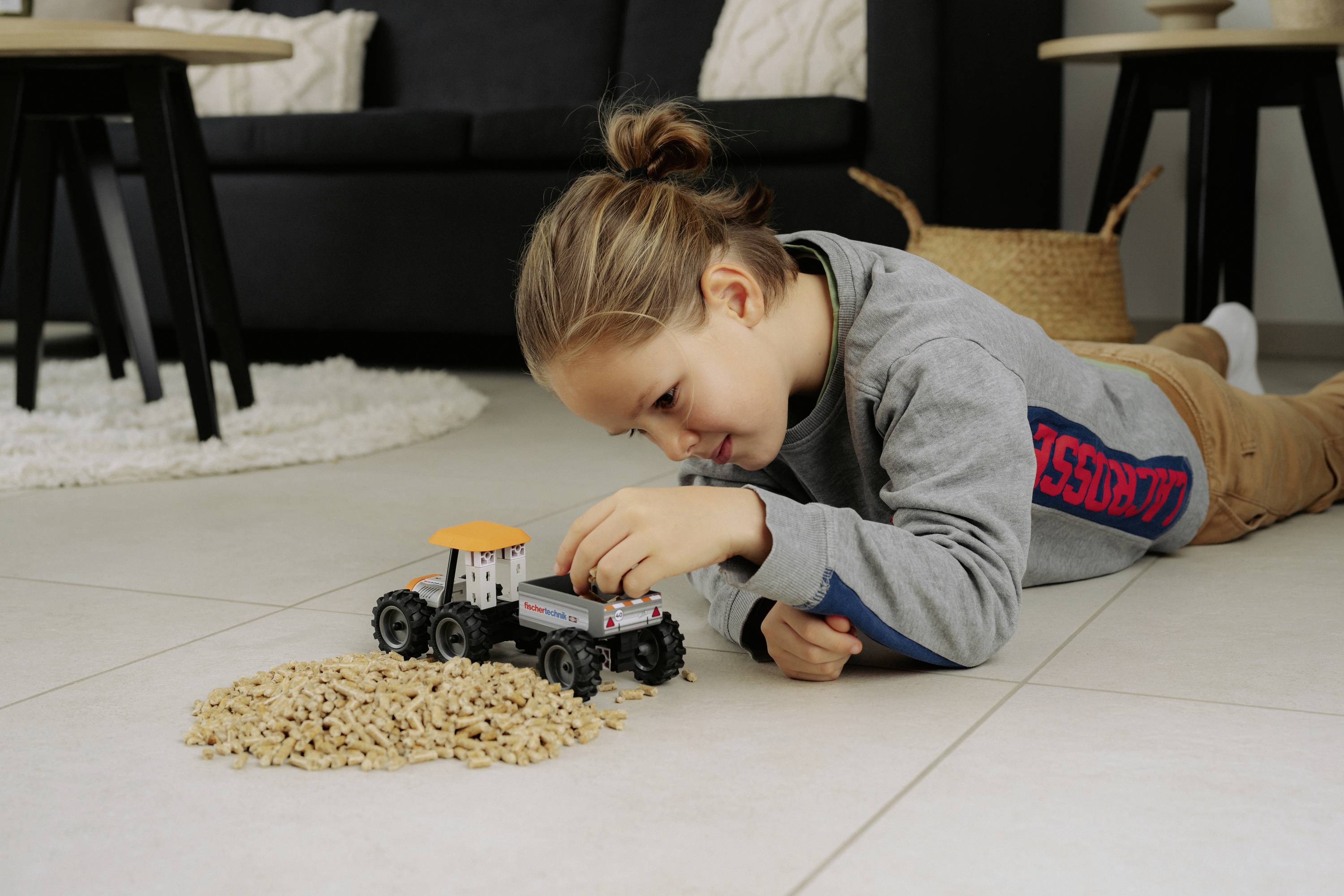 Un enfant joue sur le sol avec un tracteur miniature et un petit tas de granulés de bois, dans un salon chaleureux et douillet.