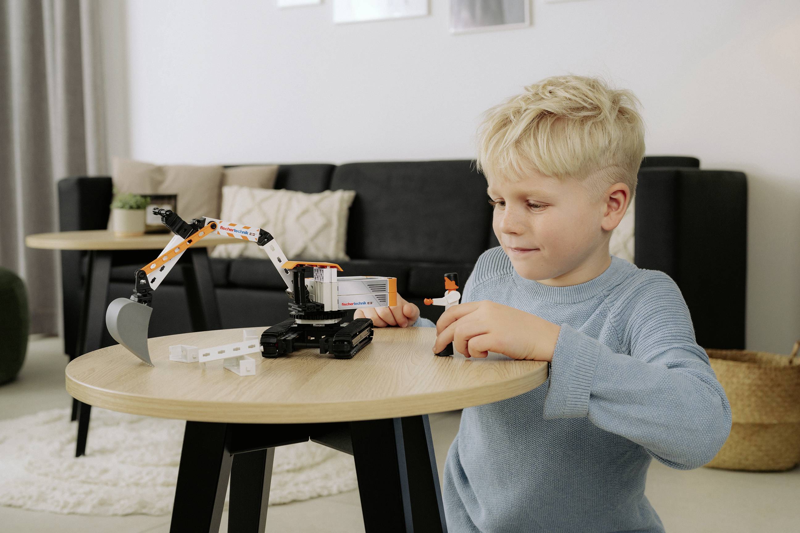Un jeune garçon portant un pull bleu assemble un véhicule de construction miniature sur une petite table en bois dans un salon.