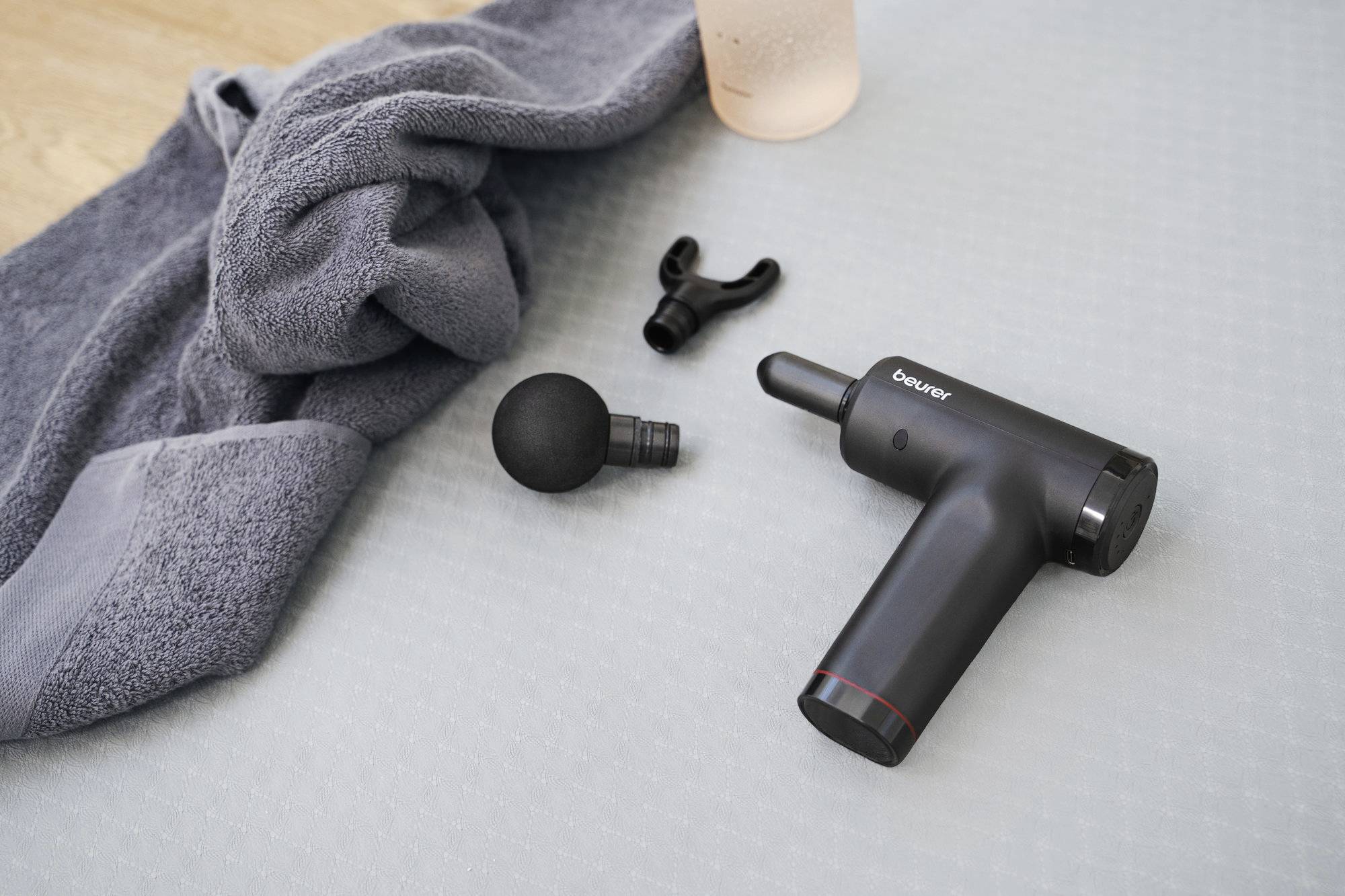 Un pistolet de massage avec deux embouts est posé sur un tapis d'exercice gris, à côté d'une serviette roulée et d'une bouteille capsée, suggérant un environnement de récupération après un entraînement.