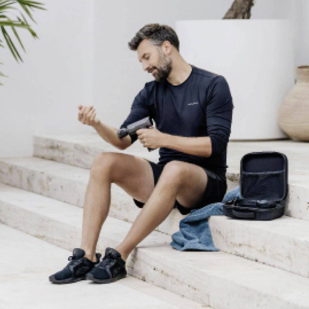 Homme assis sur des marches en pierre, utilisant un pistolet de massage sur sa jambe, avec un étui ouvert à côté de lui, portant une tenue de sport, à l'intérieur.