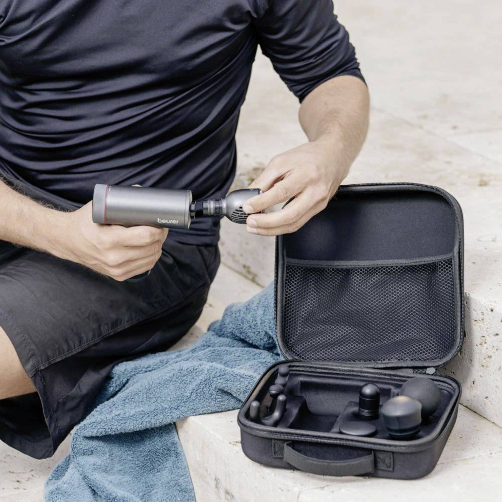 Une personne utilise un pistolet de massage sur sa jambe, avec un étui d'embouts posé au sol à côté d'elle.