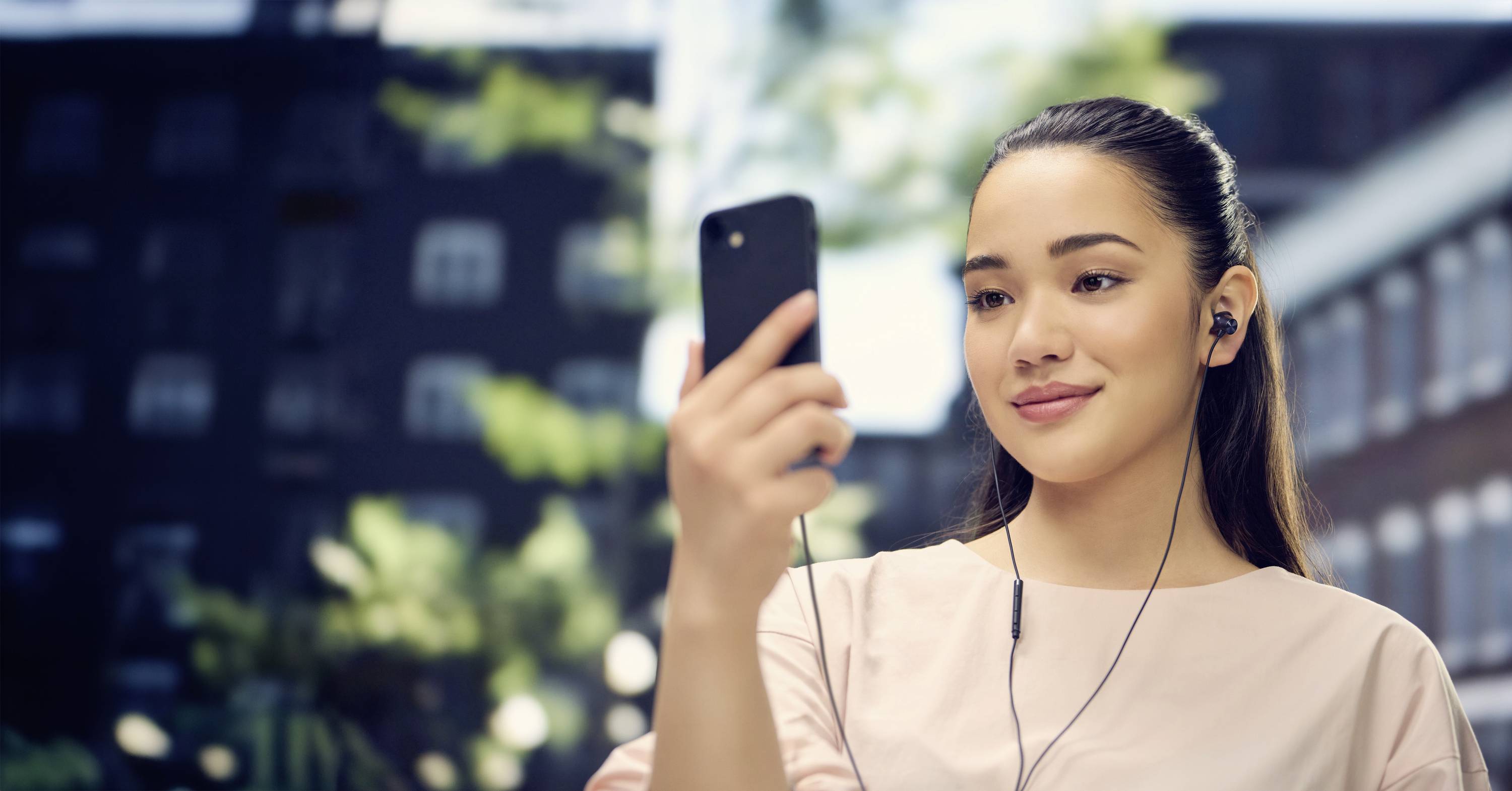 Une femme regarde son smartphone avec des écouteurs, debout dehors dans un environnement urbain par une journée ensoleillée.