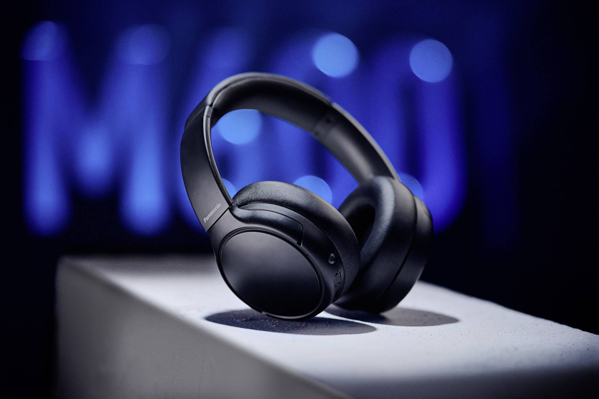 Casque audio noir supra-auriculaire posé sur un piédestal blanc, avec un arrière-plan flou bleu. L'accent est mis sur le casque.