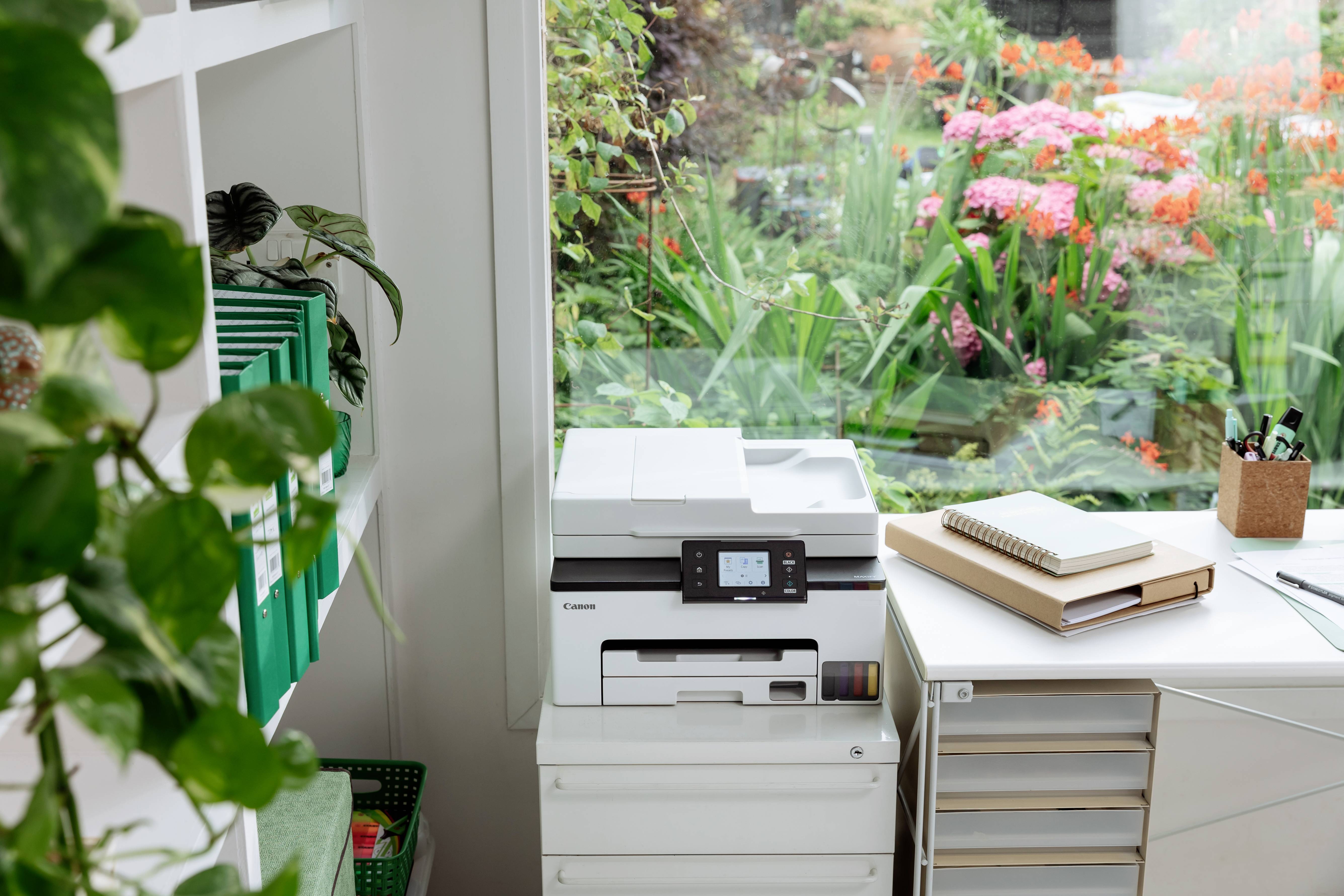 Une imprimante sur un bureau près d'une fenêtre, donnant sur un jardin aux fleurs colorées. Des étagères avec des chemises vertes et des articles de papeterie sont visibles.
