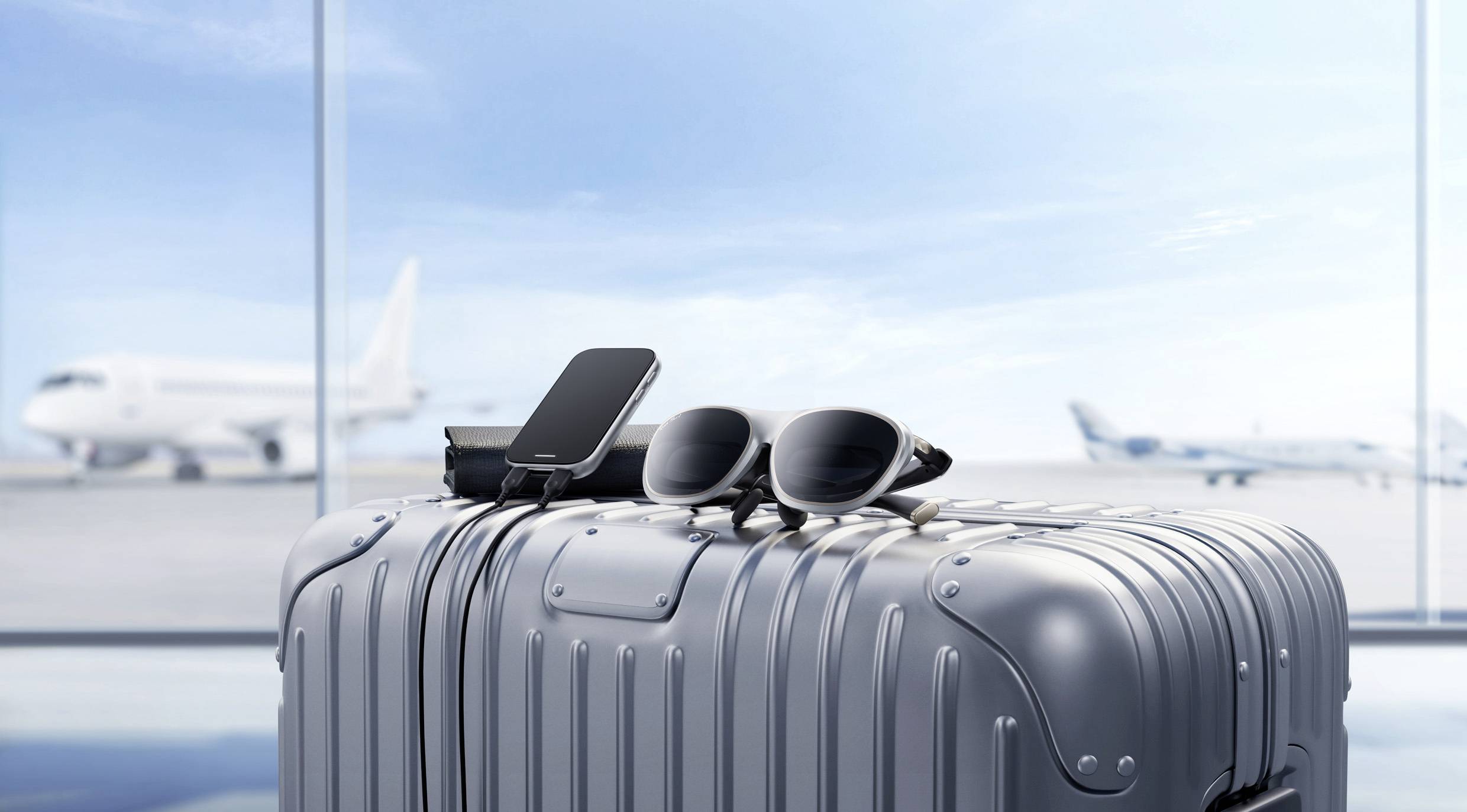 Une valise argentée dans un aéroport, avec un smartphone et des lunettes de soleil posés sur le dessus. Deux avions sont visibles à travers de grandes fenêtres en arrière-plan.