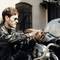 Un homme portant des lunettes de soleil et une veste en cuir répare ou ajuste une motocyclette. La scène se déroule en extérieur, à la lumière du jour, près d'un bâtiment.