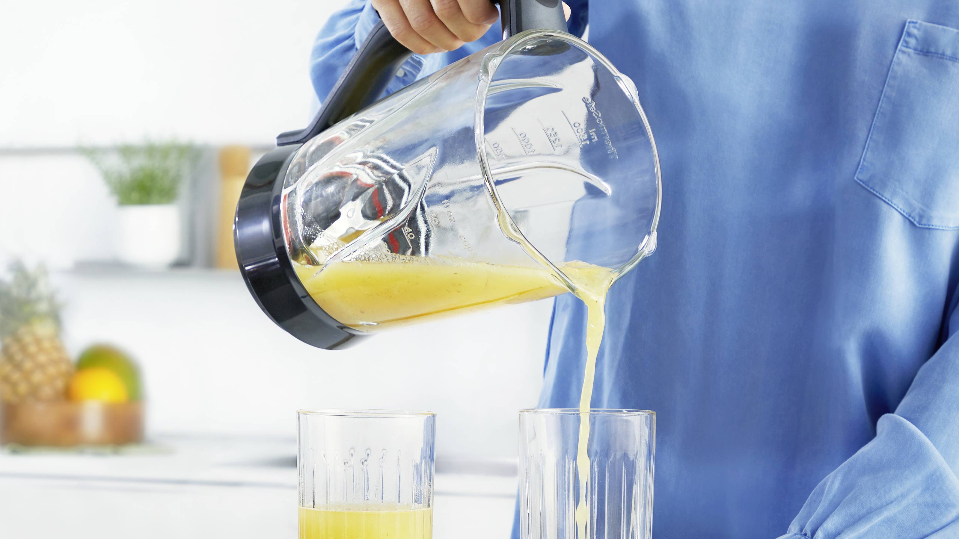 Une personne portant une chemise bleue verse du jus d'orange d'un blender dans un verre, avec des accessoires de cuisine et un bol de fruits en arrière-plan.