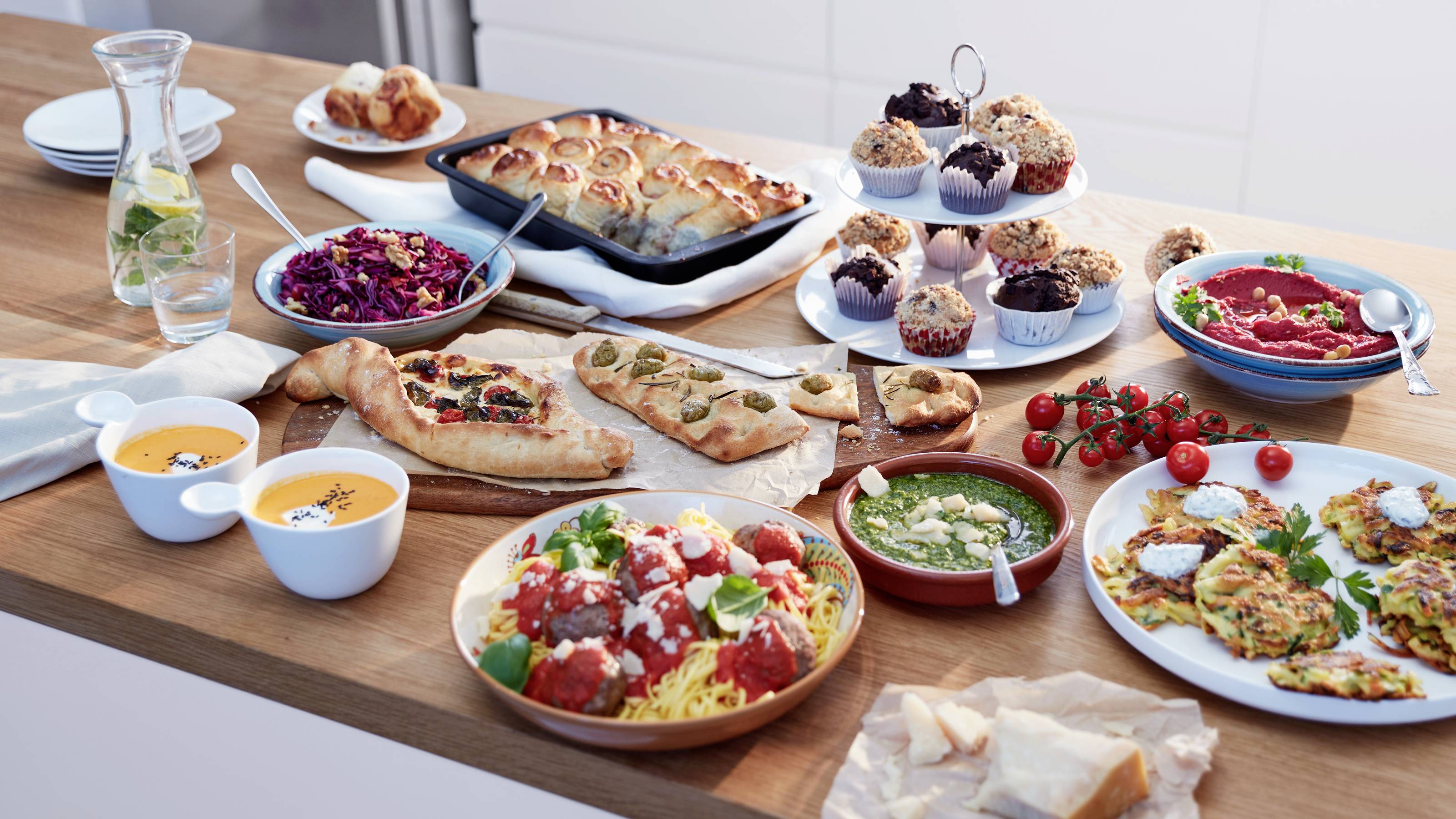 Une table en bois garnie de plats variés, comprenant des cupcakes, des salades, des soupes, des pâtes aux boulettes de viande et du pain, disposés pour un buffet.