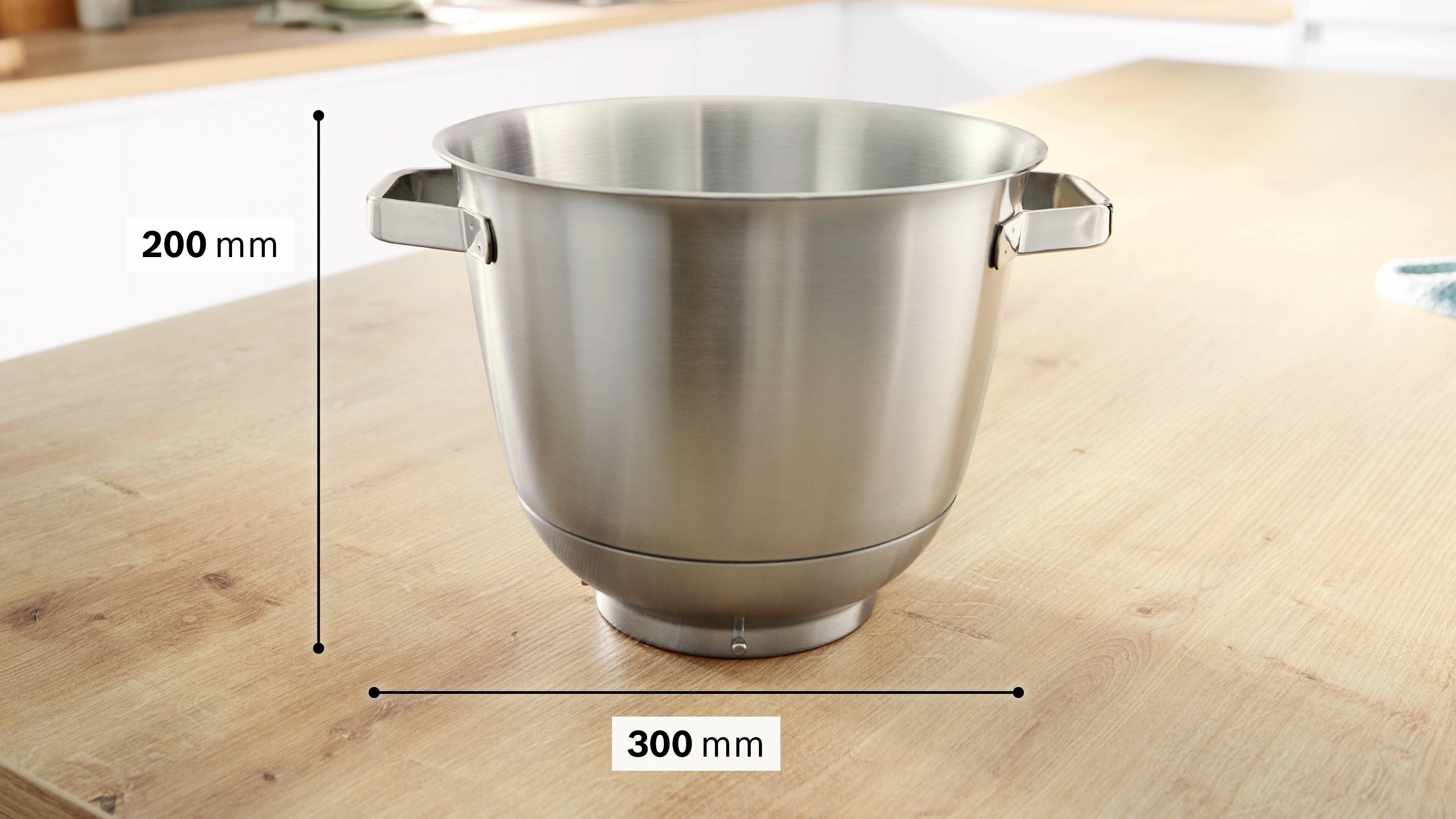 Un bol à mélanger en métal posé sur un plan de travail en bois, haut de 200 mm et large de 300 mm, équipé de deux petites poignées sur les côtés.