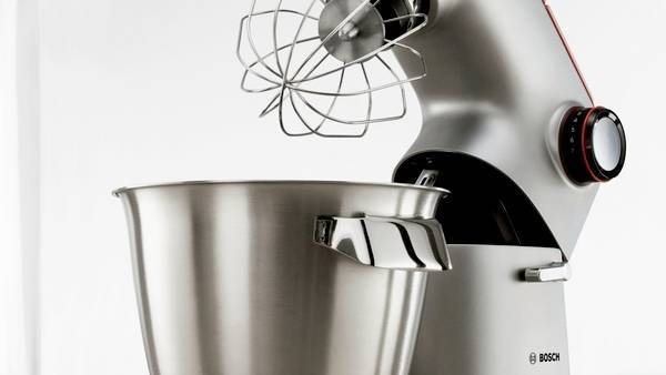 Un robot pâtissier argenté avec son fouet est placé au-dessus d'un grand bol en métal, prêt à être utilisé dans un environnement de cuisine.