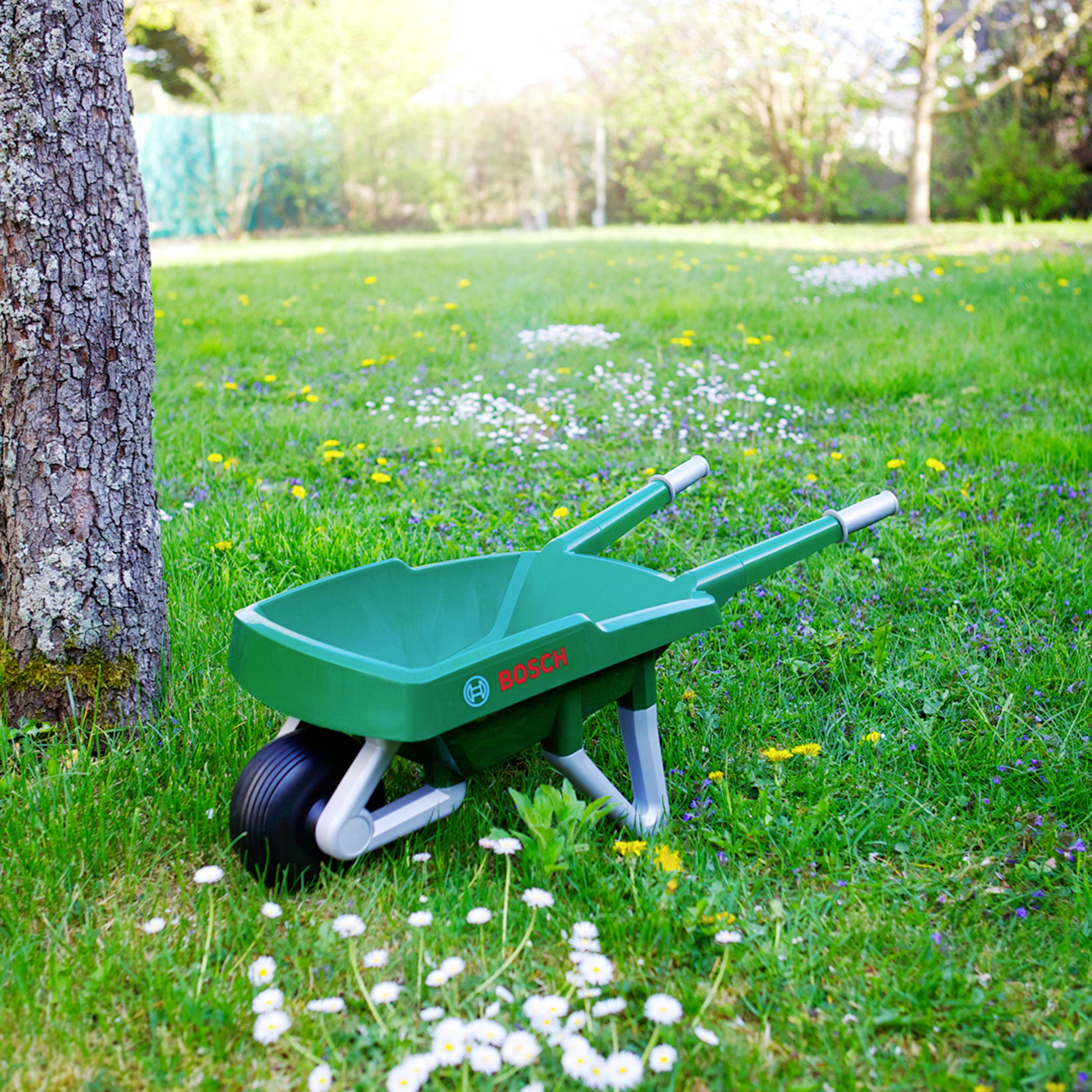 Une brouette verte pour enfants avec la marque 'Bosch' est positionnée verticalement sur une pelouse herbeuse près d'un arbre, entourée de marguerites éparses.