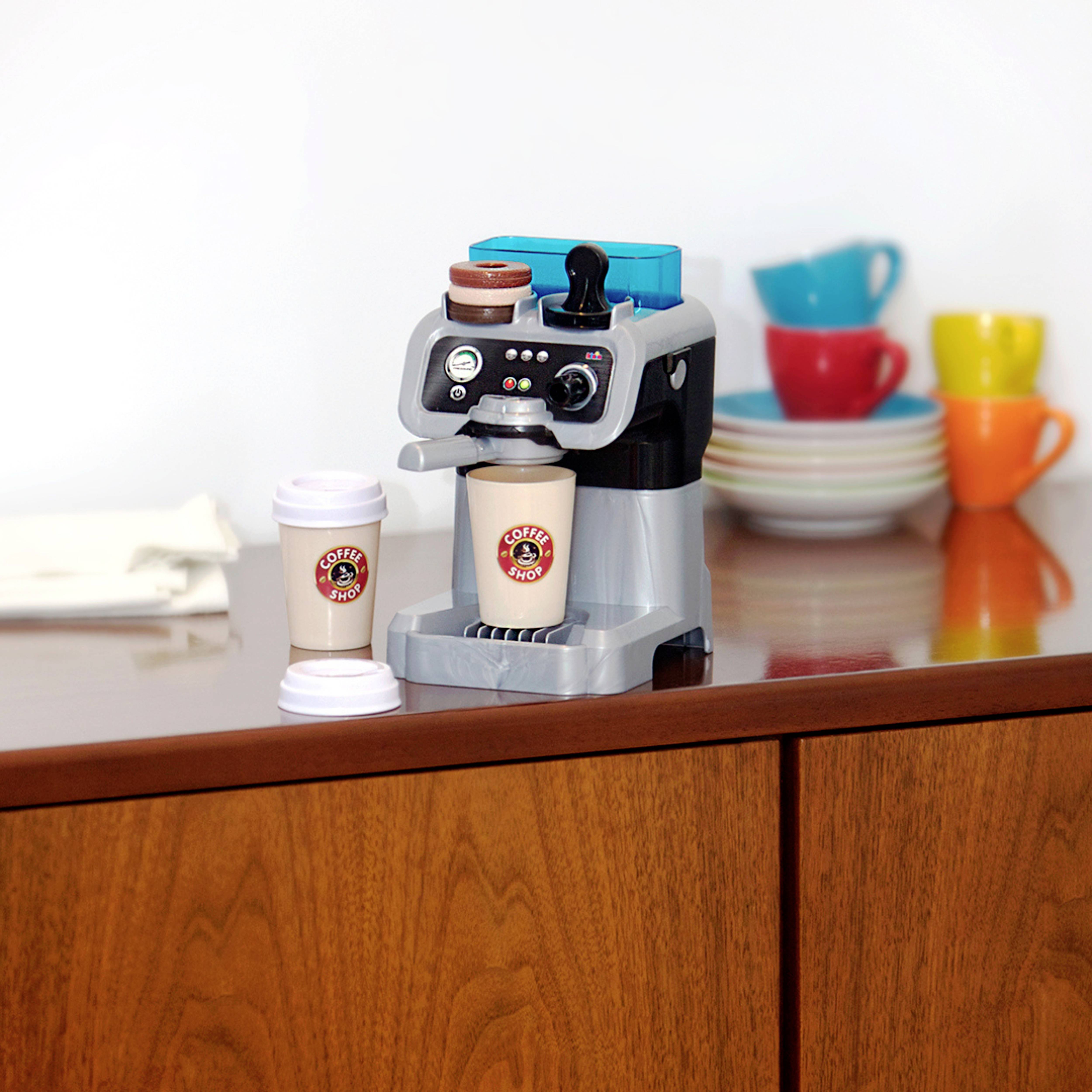 Un petit cafetière jouet avec deux tasses de café jouets étiquetées 'Coffee Shop' sur un comptoir en bois, avec des assiettes et des mugs colorés empilés en arrière-plan.