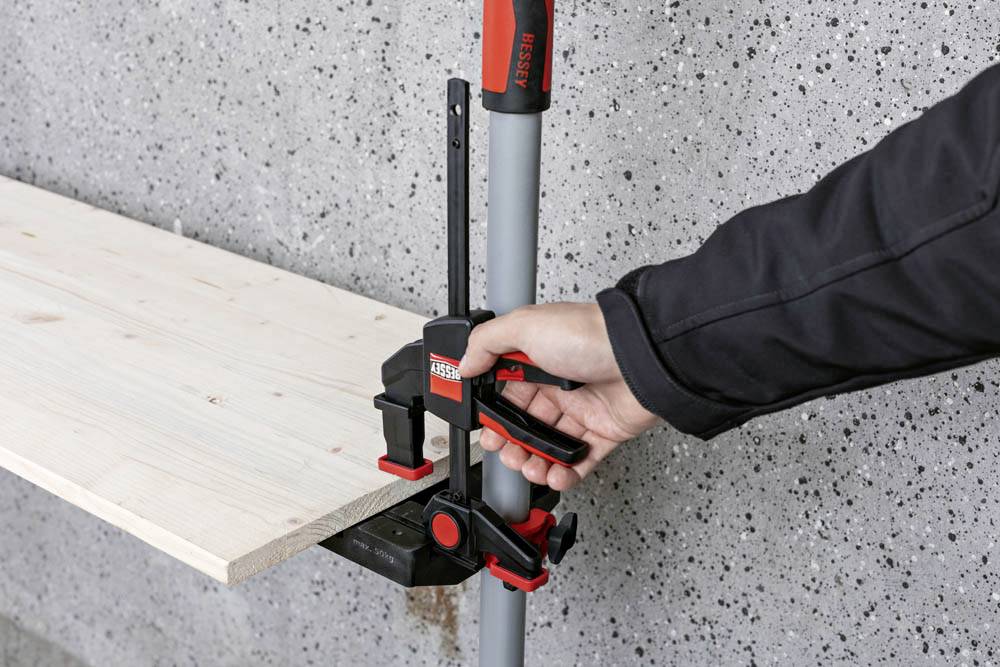 Une personne resserre un collier de serrage sur un tuyau vertical, maintenant une planche de bois contre un mur en béton. Le collier est rouge, noir et gris.