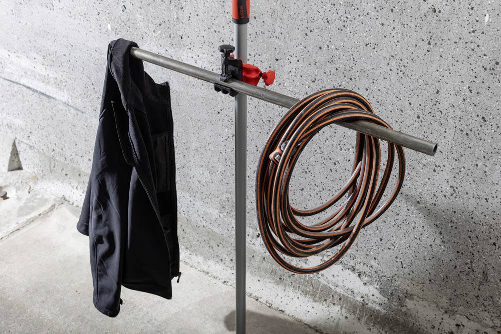 Un support métallique maintient une veste noire et un tuyau orange enroulé, disposés contre un mur en béton dans un garage ou un espace industriel.
