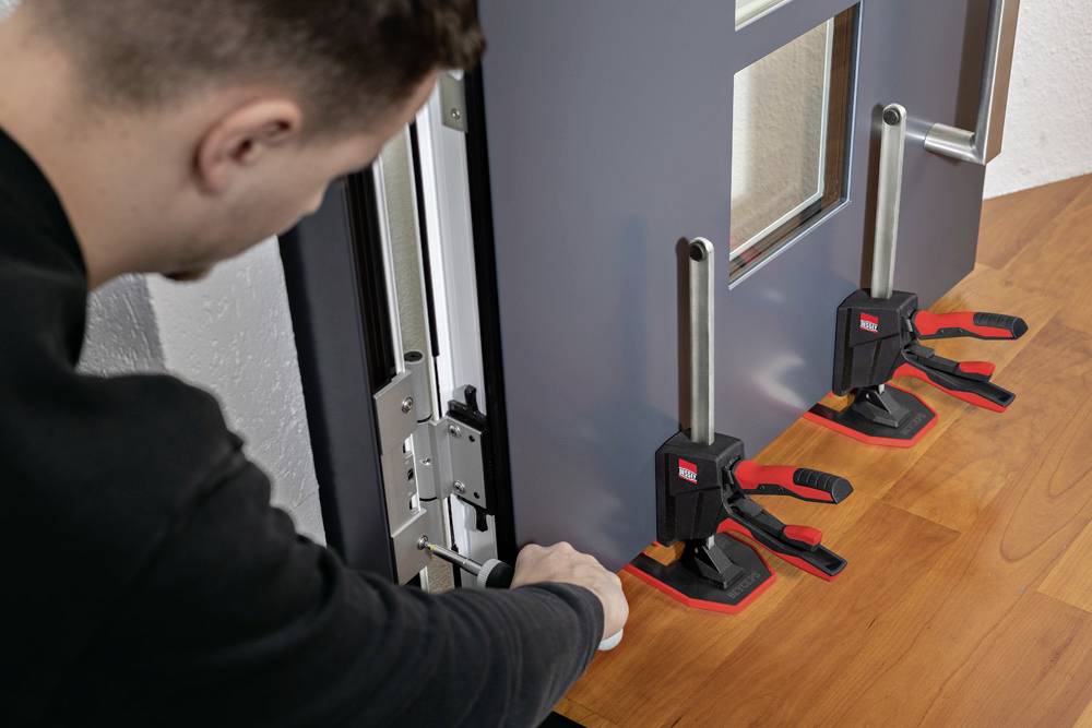 Une personne ajuste deux pinces maintenant le bord d'une porte sur une table, en se concentrant sur un alignement précis.