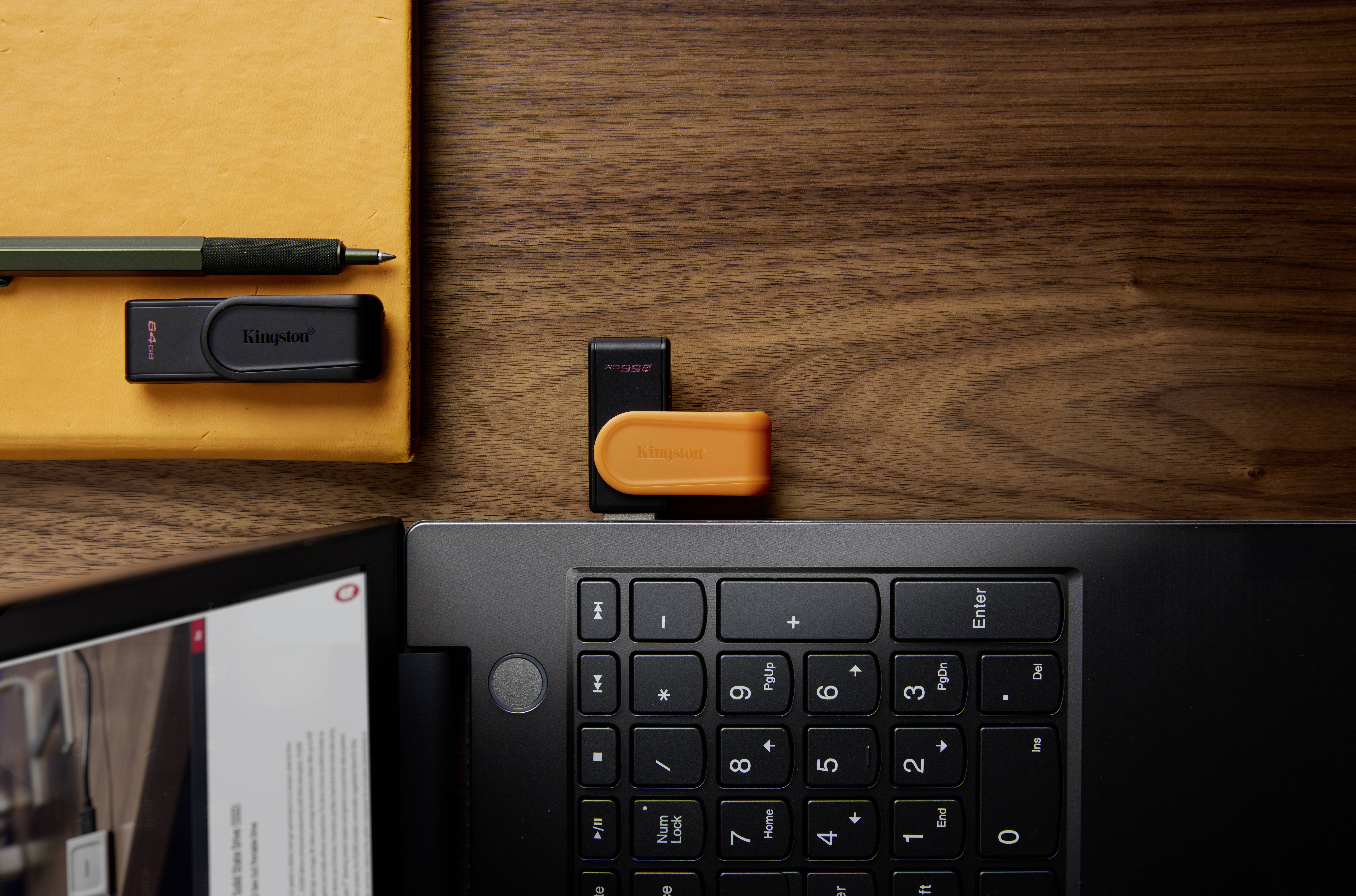 Un ordinateur portable posé sur une table en bois avec une clé USB insérée. À proximité se trouvent un carnet orange, un stylo et une autre clé USB.