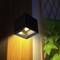 Un luminaire mural moderne au design carré éclaire une paroi en bois, projetant une lumière douce sur la verdure environnante.