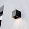 Un luminaire mural cubique noir élégant diffuse une lumière douce sur un mur gris texturé, créant une esthétique moderne et minimaliste.