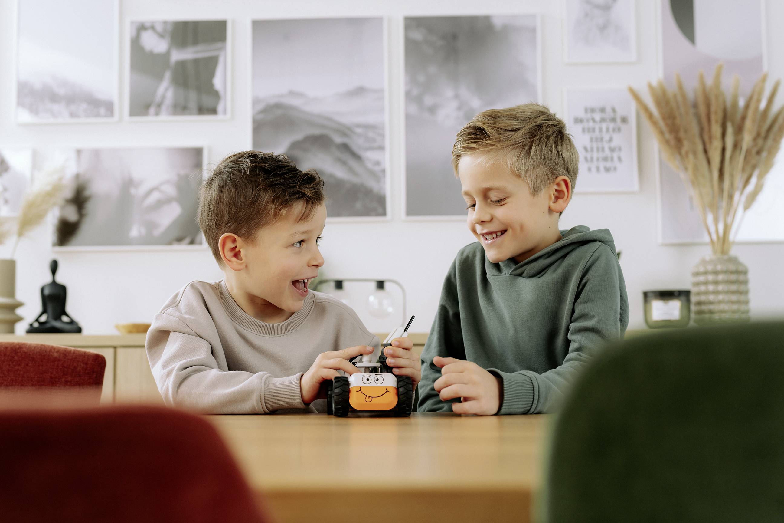 Deux jeunes garçons sont assis à une table, souriant et jouant avec un petit robot jouet. Des œuvres encadrées décorent le mur en arrière-plan.