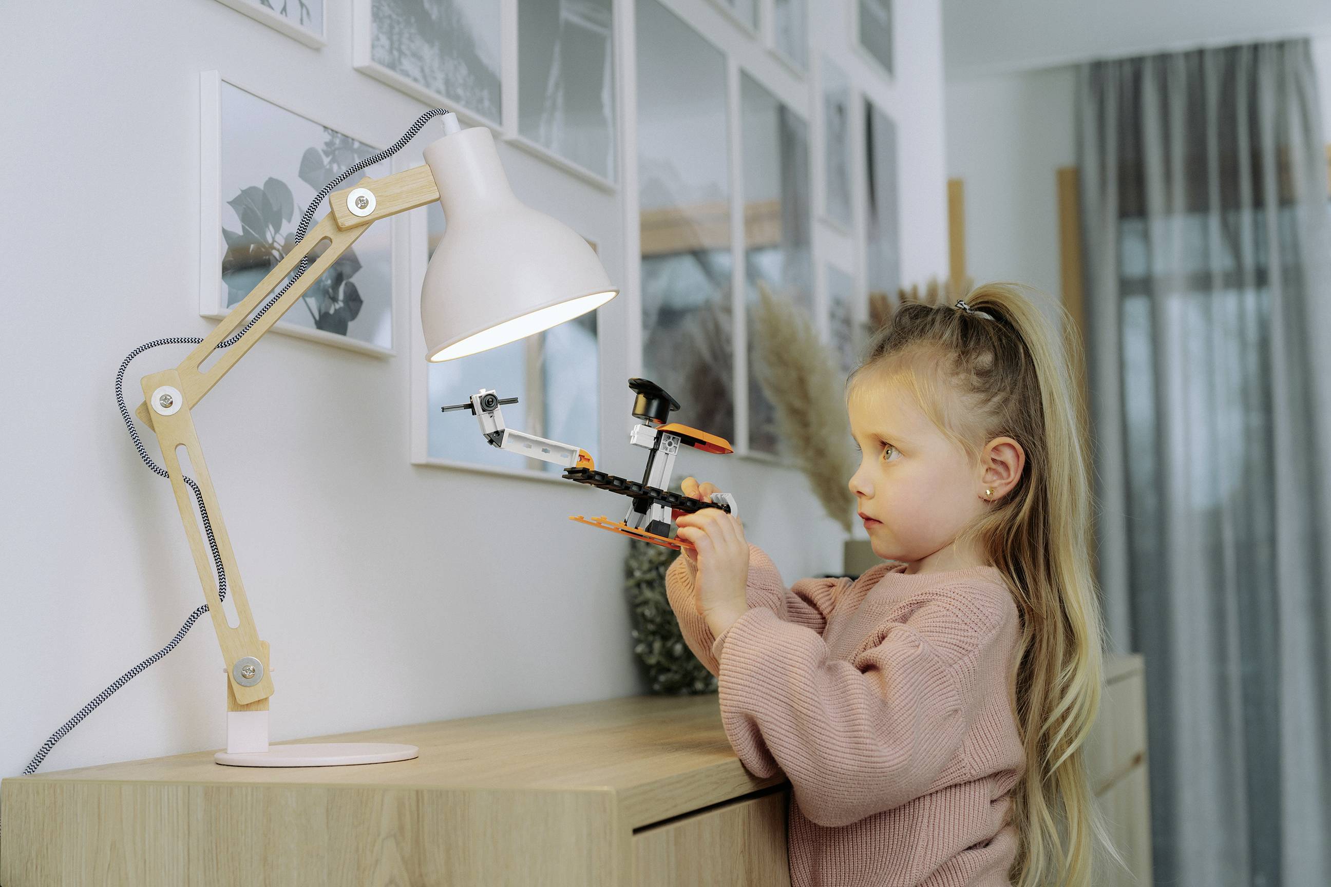 Une fille portant un pull rose assemble un jouet modèle ressemblant à un drone sur un bureau en bois sous une lampe de bureau blanche dans un environnement domestique.
