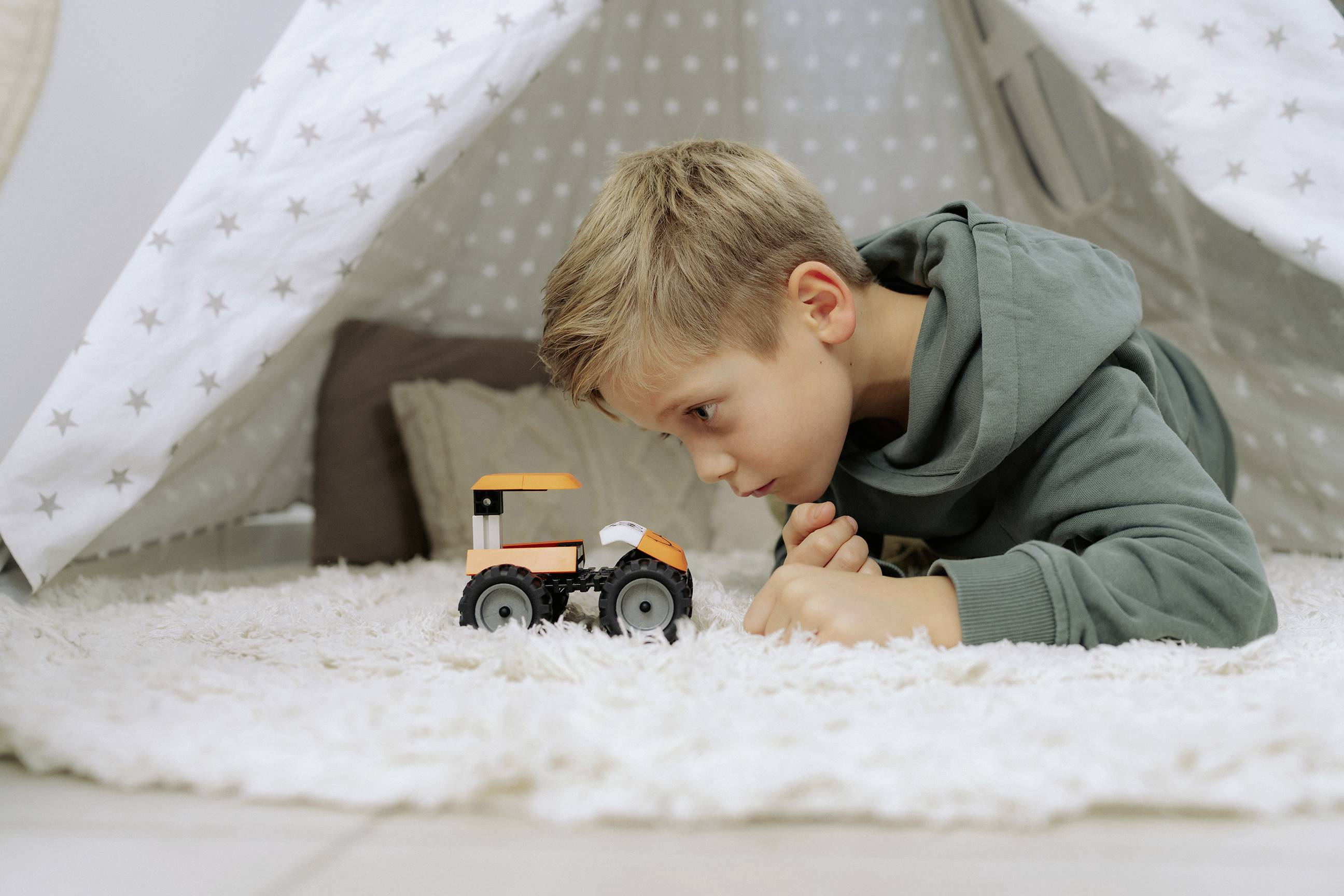 Un enfant en sweat à capuche vert est allongé dans une cabane de couvertures, concentré intensément sur un petit tracteur jouet posé sur le tapis.