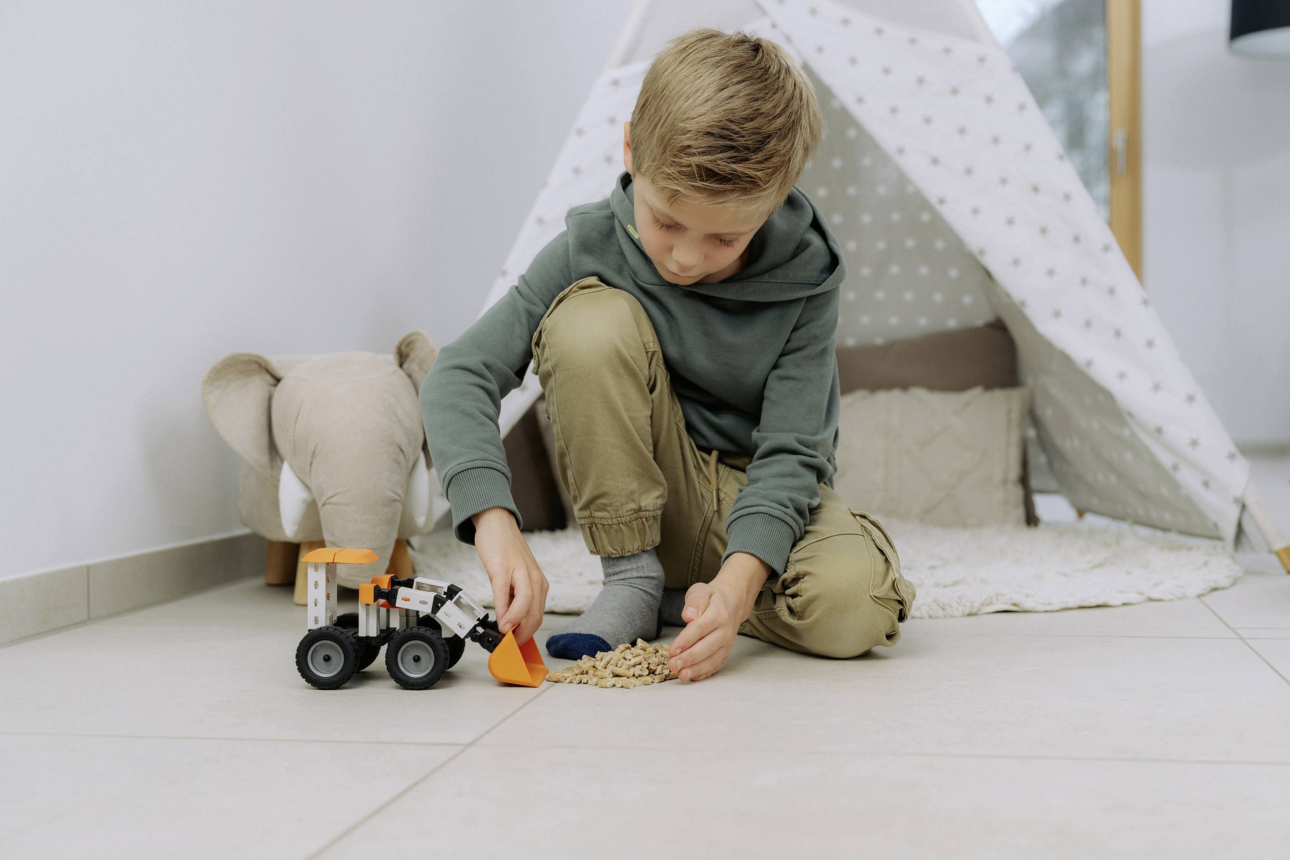 Un enfant joue sur le sol d'une pièce confortable, ramassant des céréales avec un chargeur frontal jouet. Une tente et un éléphant en peluche sont en arrière-plan.