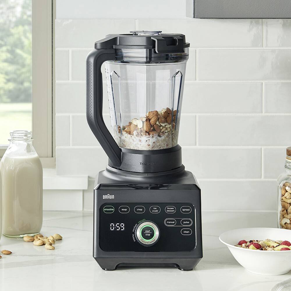 Un mixeur sur un plan de travail de cuisine rempli d'amandes et de flocons d'avoine. À proximité se trouvent une bouteille de lait et un bocal, suggérant la préparation d'un smoothie.