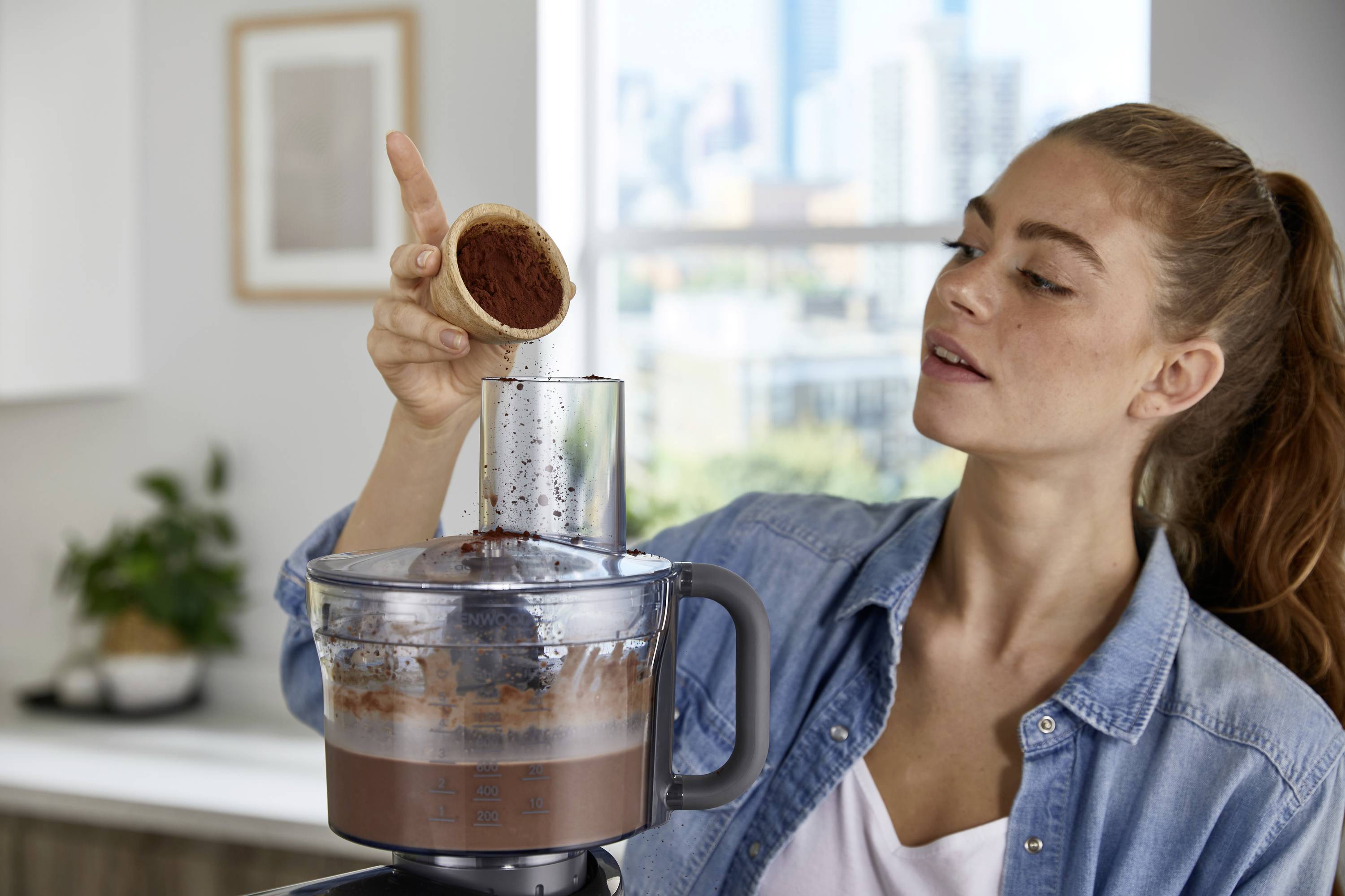 Une femme ajoute de la poudre de cacao à un robot culinaire contenant de la pâte à chocolat, dans une cuisine moderne avec un paysage urbain flou en arrière-plan.