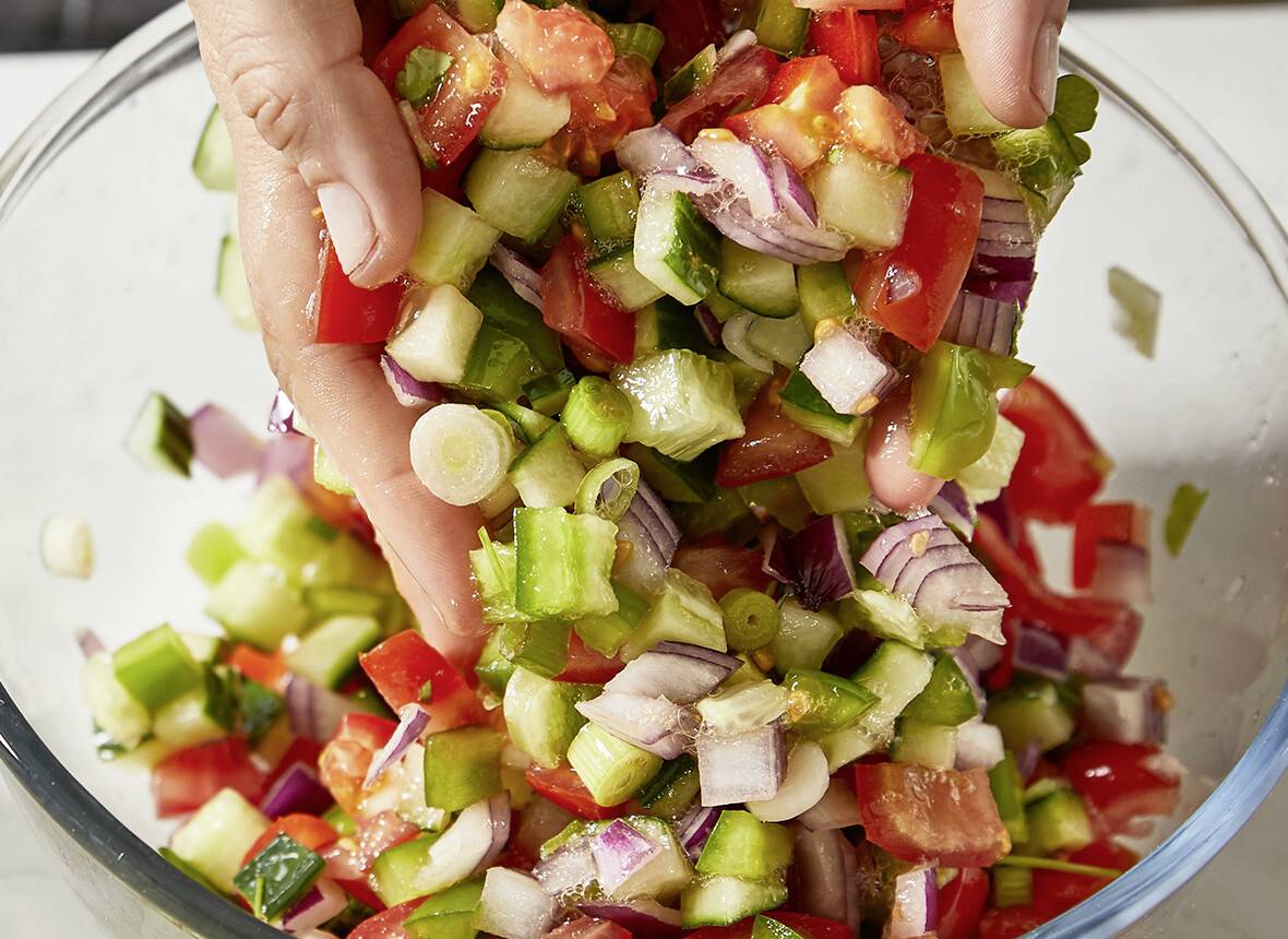 Des mains mélangeant un assortiment coloré de légumes hachés comprenant des poivrons, des oignons et des concombres dans un bol transparent.