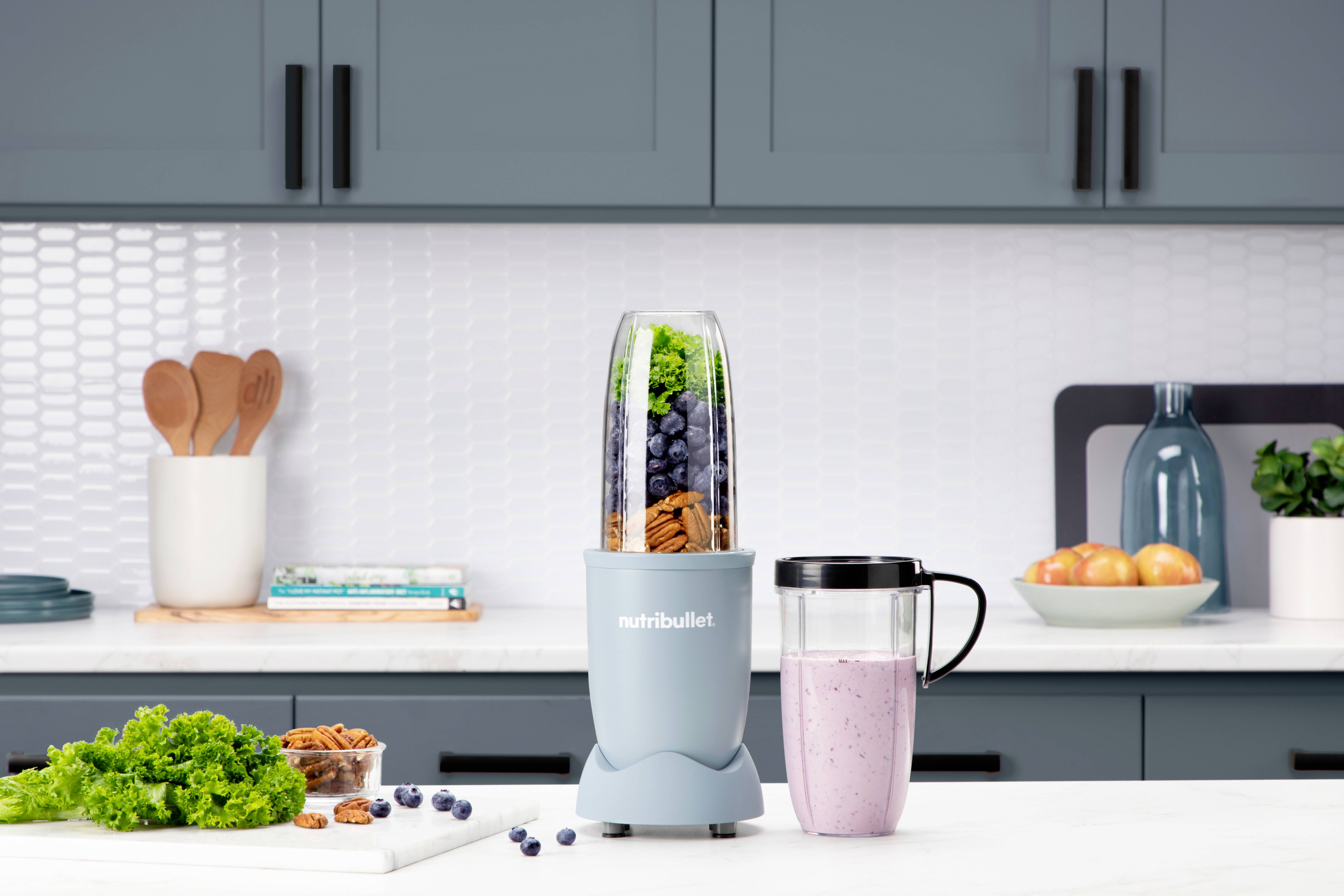 Un comptoir de cuisine avec un blender à smoothies rempli de légumes verts, de baies et de noix. Une tasse de smoothie et divers ustensiles de cuisine sont en arrière-plan.