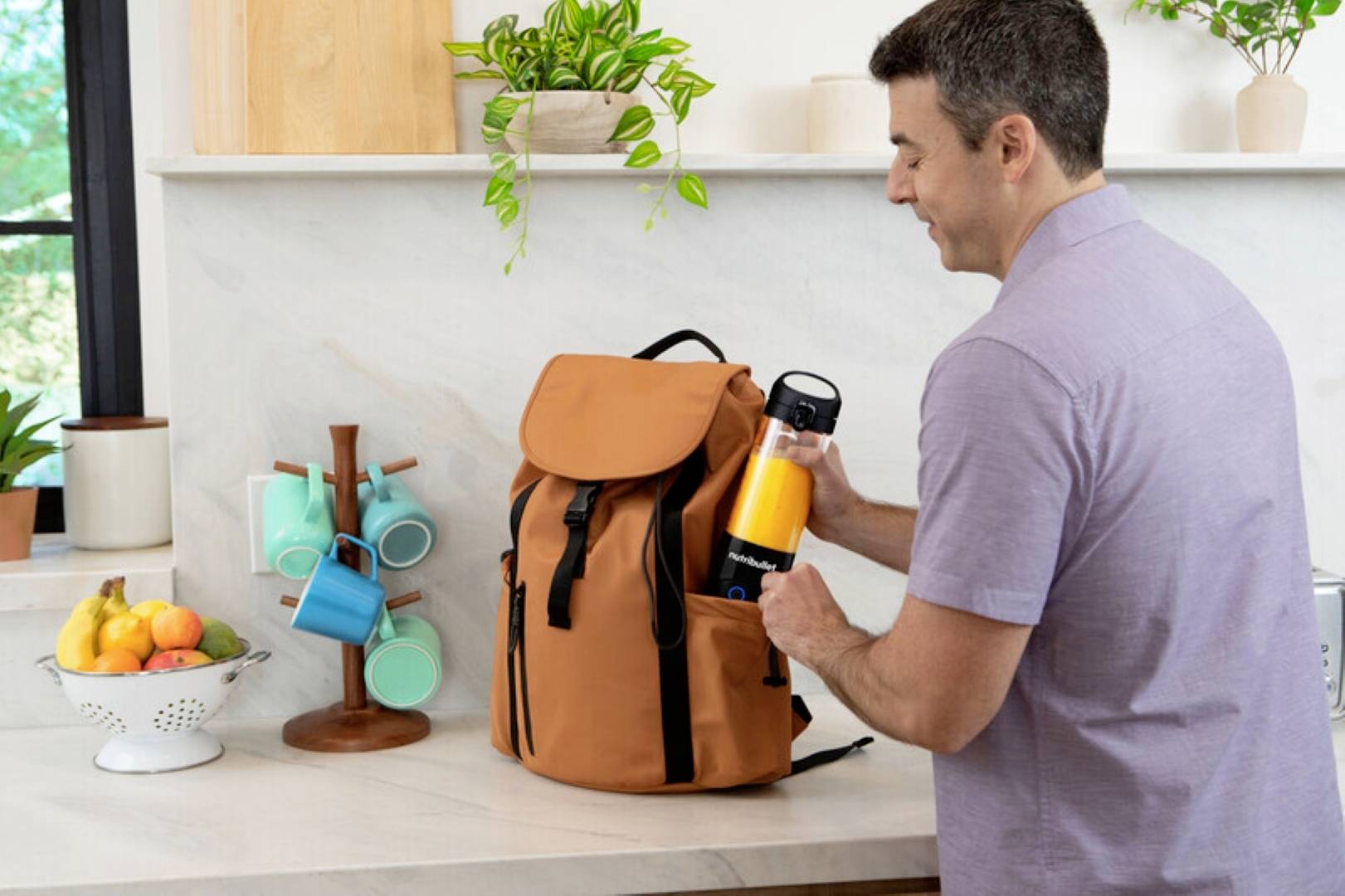 Un homme place une bouteille d'eau dans un sac à dos marron posé sur un comptoir de cuisine. À proximité, des tasses sont empilées et un bol de fruits est présent.
