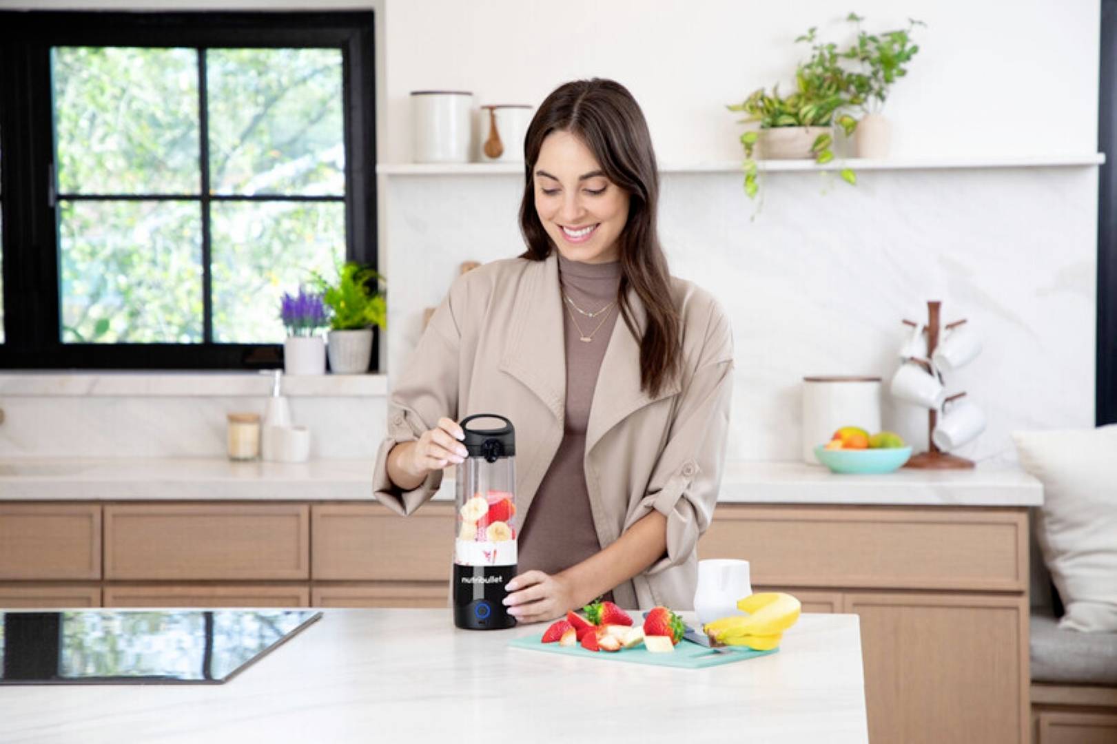 Une femme dans une cuisine mixe des fruits avec un mixeur personnel. Des fruits frais sont posés sur le comptoir, et la cuisine présente une décoration moderne.