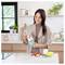 Une femme dans une cuisine prépare un smoothie avec un blender. À proximité, des bananes et des fraises sont posées sur une planche à découper. La cuisine est lumineuse et moderne.
