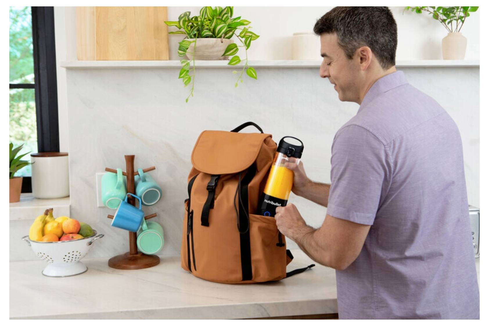 Un homme range une bouteille de jus d'orange dans un sac à dos brun sur un comptoir de cuisine, avec des fruits et des gobelets, suggérant une préparation pour une sortie.