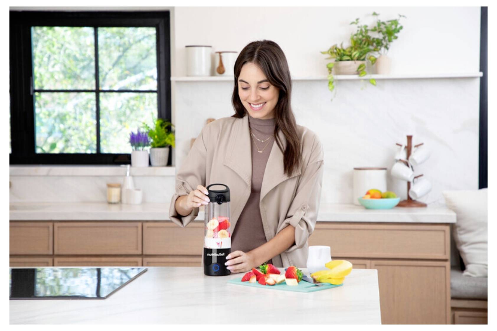 Une personne dans une cuisine prépare un smoothie avec un blender portable, entourée de fruits coupés sur le comptoir, suggérant la préparation d'un repas frais.