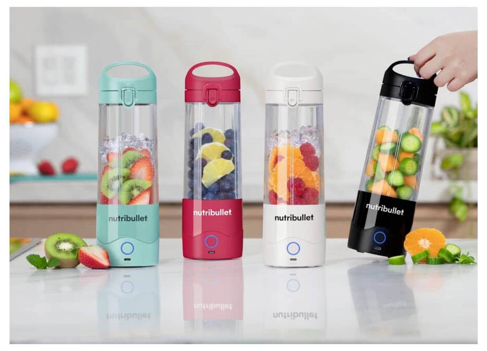 Quatre blenders NutriBullet de différentes couleurs, chacun rempli de fruits et de glace, sont posés sur un plan de travail. Une main tient le blender noir.