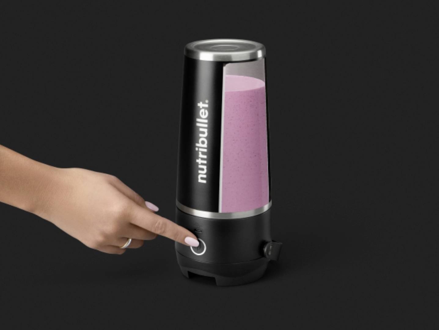 La main d'une personne appuie sur le bouton d'un blender Nutribullet contenant un smoothie rose sur un fond noir.