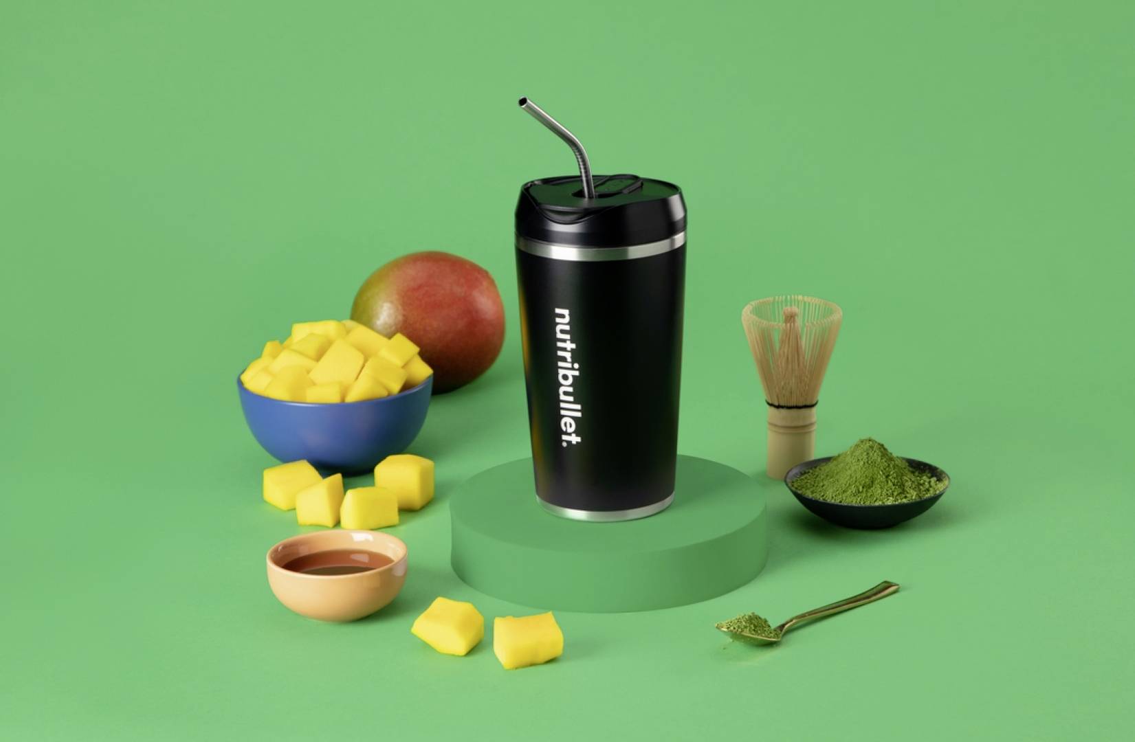 Un gobelet Nutribullet noir avec une paille sur une plateforme verte, entouré de mangue en dés, d'une mangue entière, de poudre de matcha et d'un fouet.