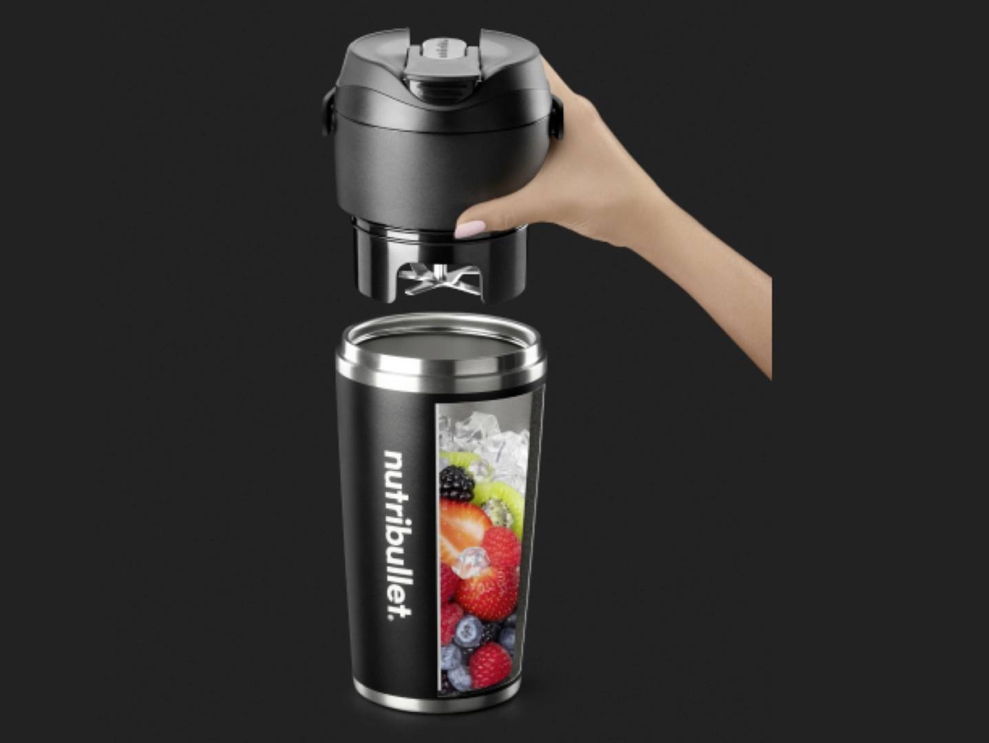 Un gobelet noir de blender NutriBullet rempli de glaçons et de fruits variés est placé sur sa base motorisée noire par une main.