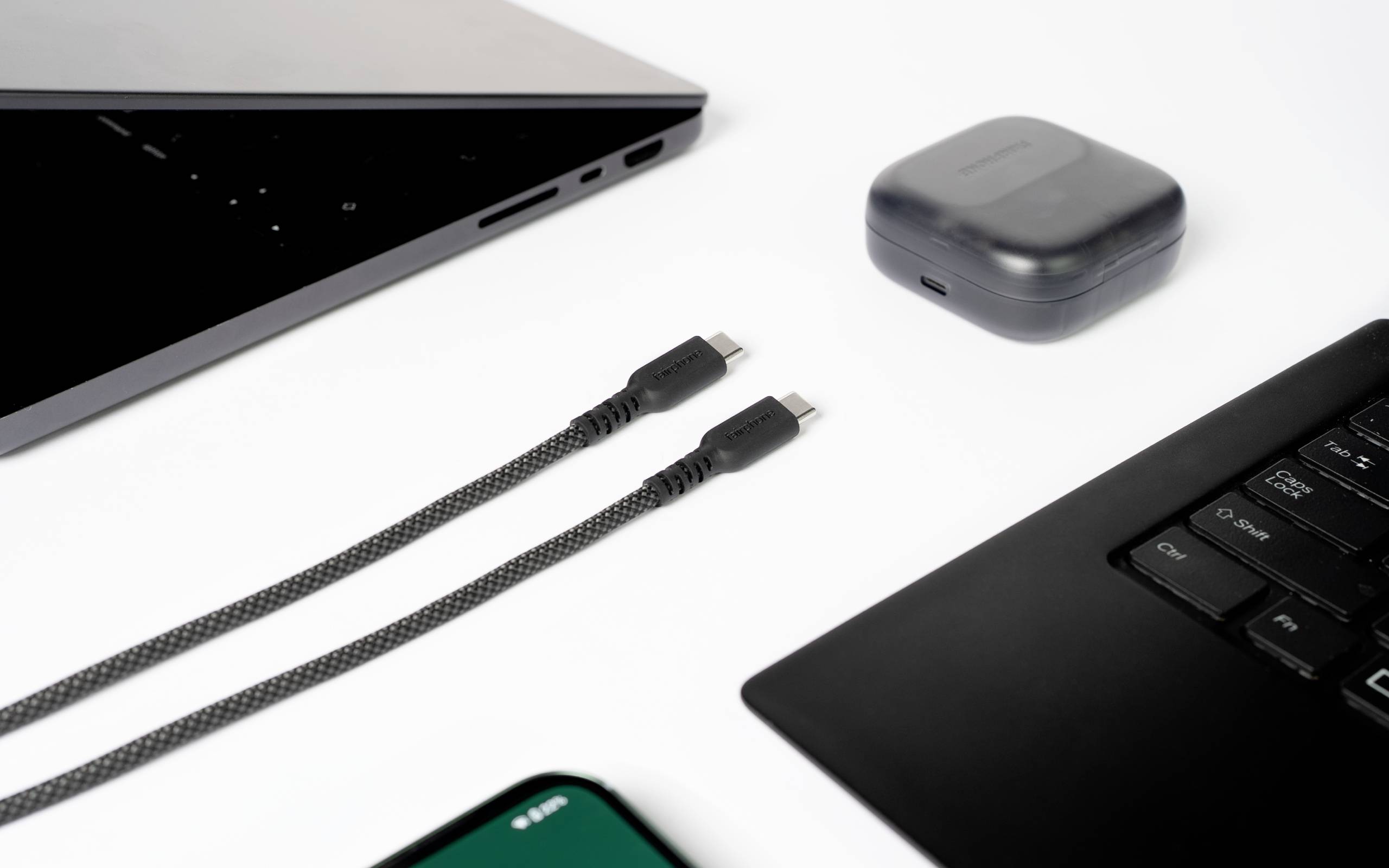 Un câble USB-C noir est placé entre un ordinateur portable, un boîtier d'écouteurs et une partie d'un smartphone, tous sur une surface blanche.