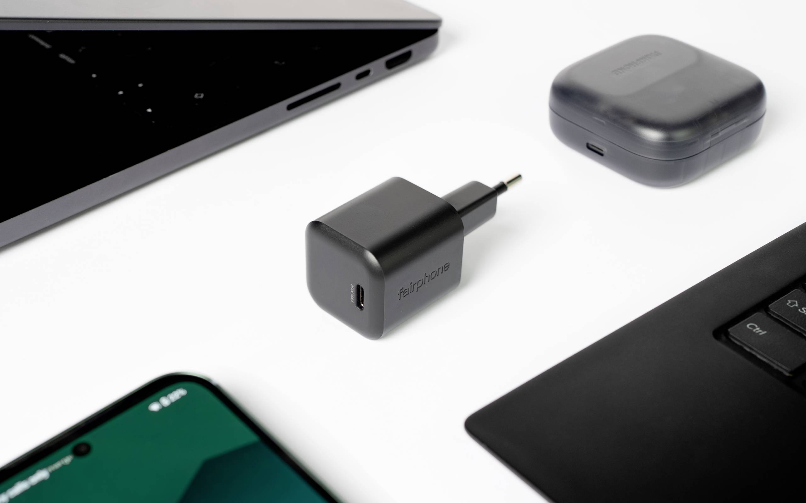 Un chargeur USB noir est posé sur une surface blanche, entouré d'un smartphone, d'un ordinateur portable et d'un étui pour écouteurs sans fil, illustrant un environnement technologique.