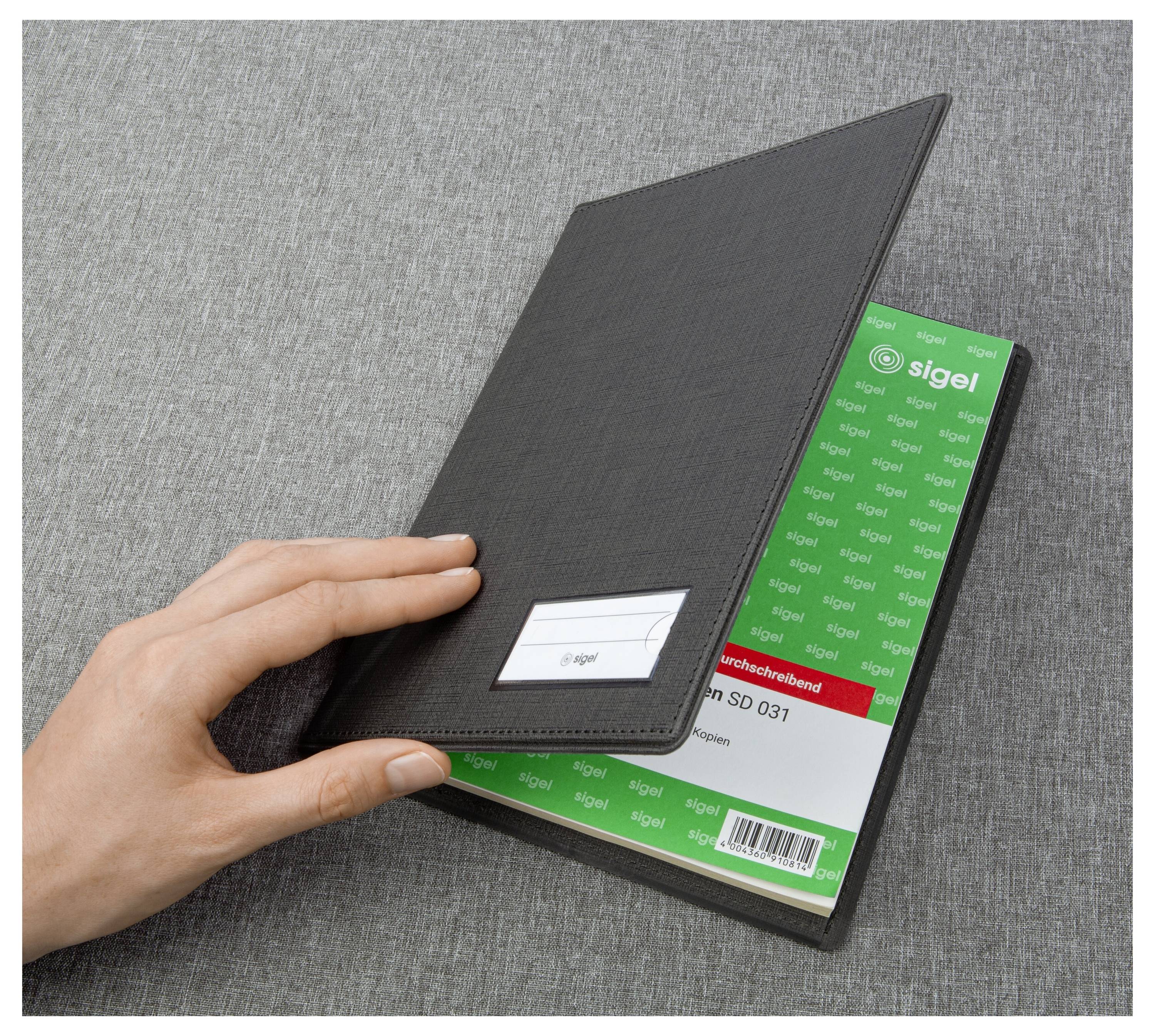 Main tenant un portefeuille en cuir noir au-dessus d'un bloc-notes ou d'un bloc de documents vert « Sigel » sur une surface en tissu gris.