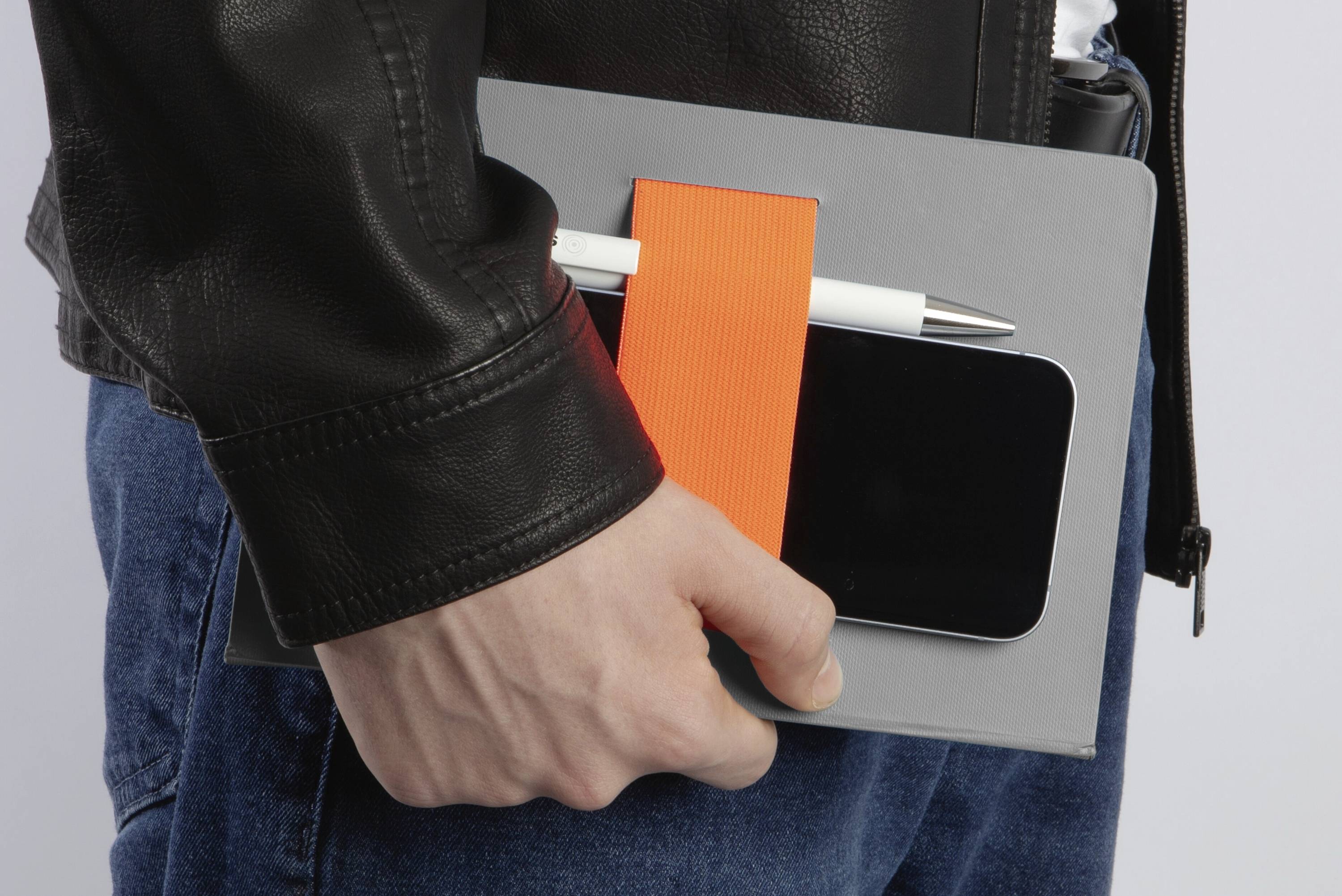 Une personne portant une veste en cuir tient une tablette grise sécurisée par une bande orange, avec un stylo et un smartphone rangés en dessous.