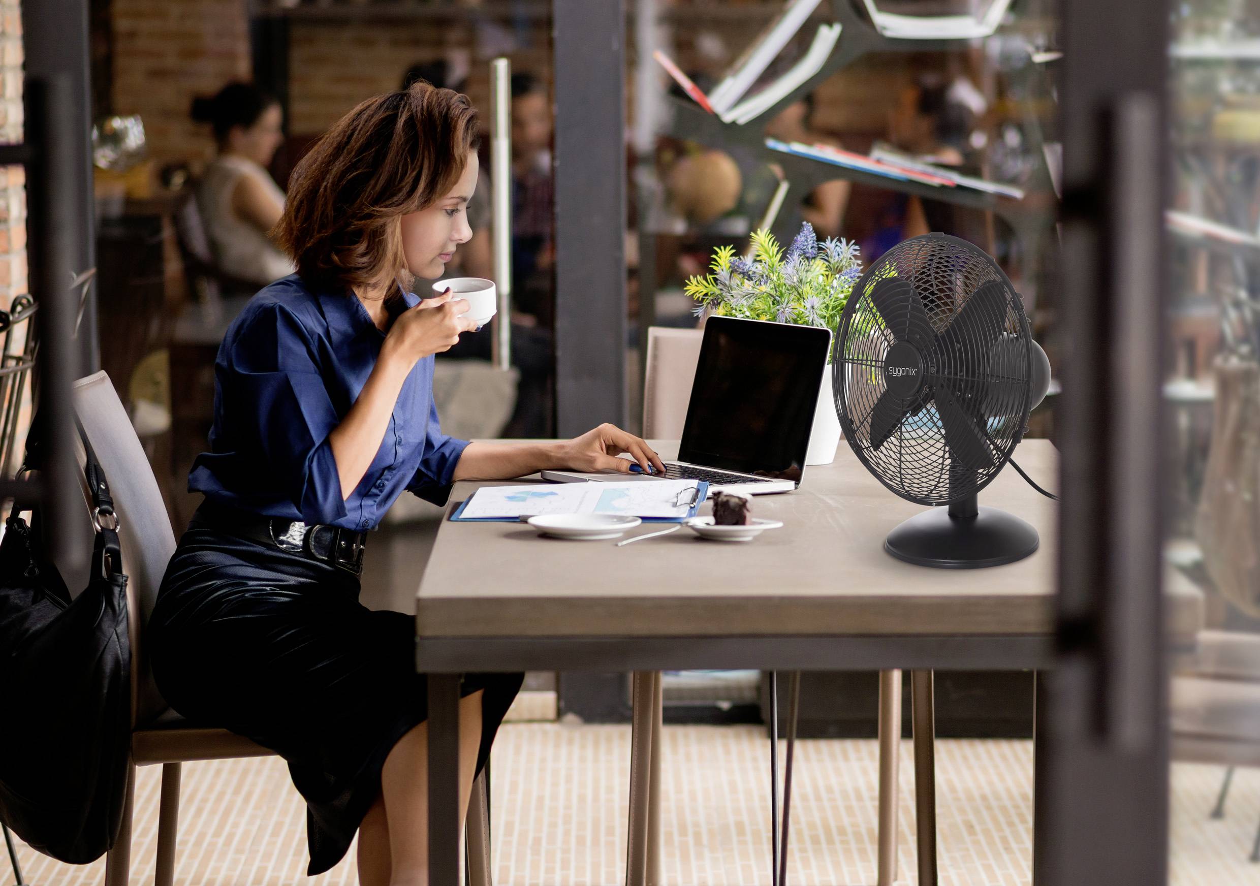 Une femme est assise à une table de café, sirotant un café et travaillant sur un ordinateur portable. Un ventilateur et une petite plante sont posés sur la table. Ambiance intérieure et détendue.